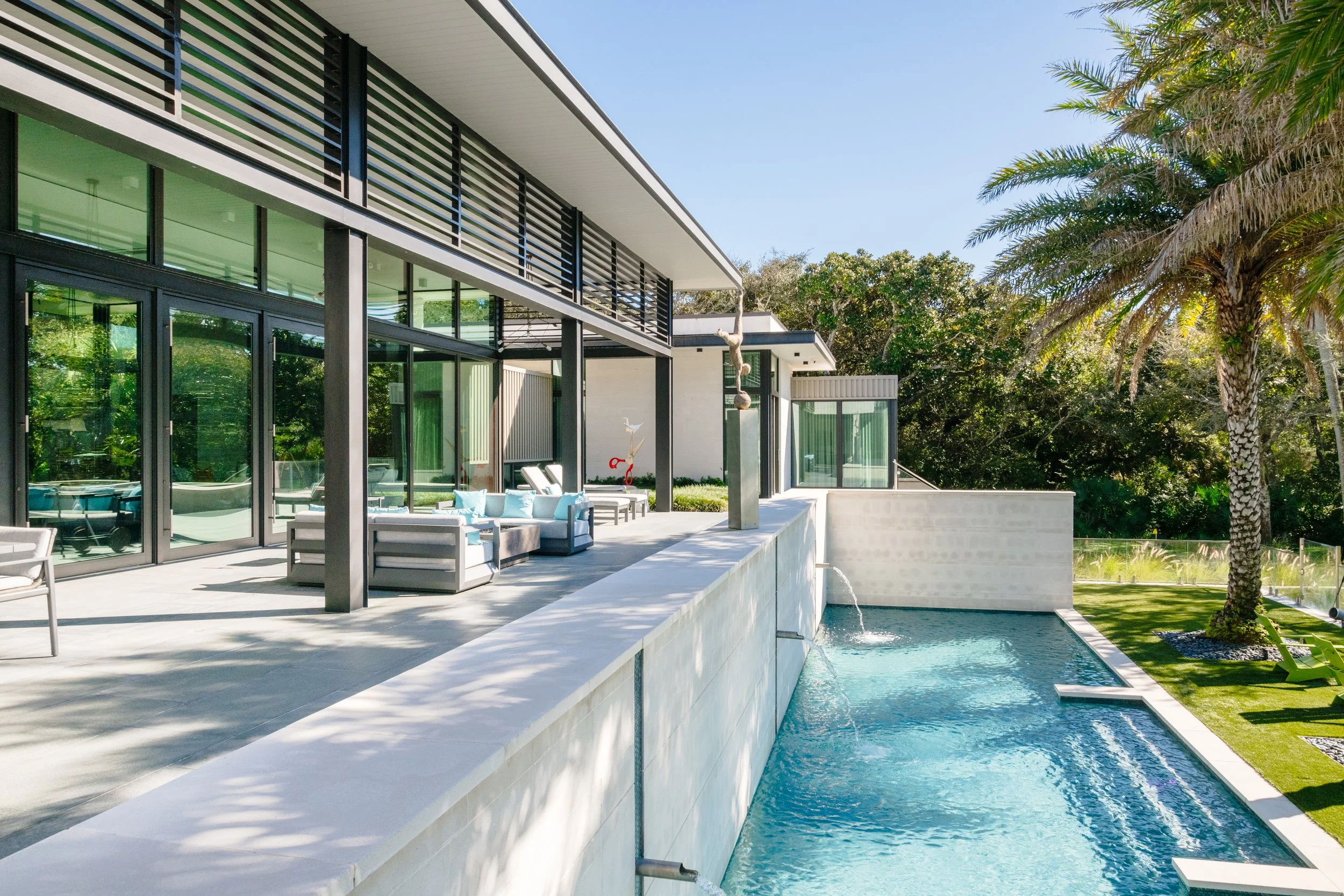Modern house with large glass windows and sliding doors, outdoor patio with seating, a pool, and lush green trees and palm trees in the background.