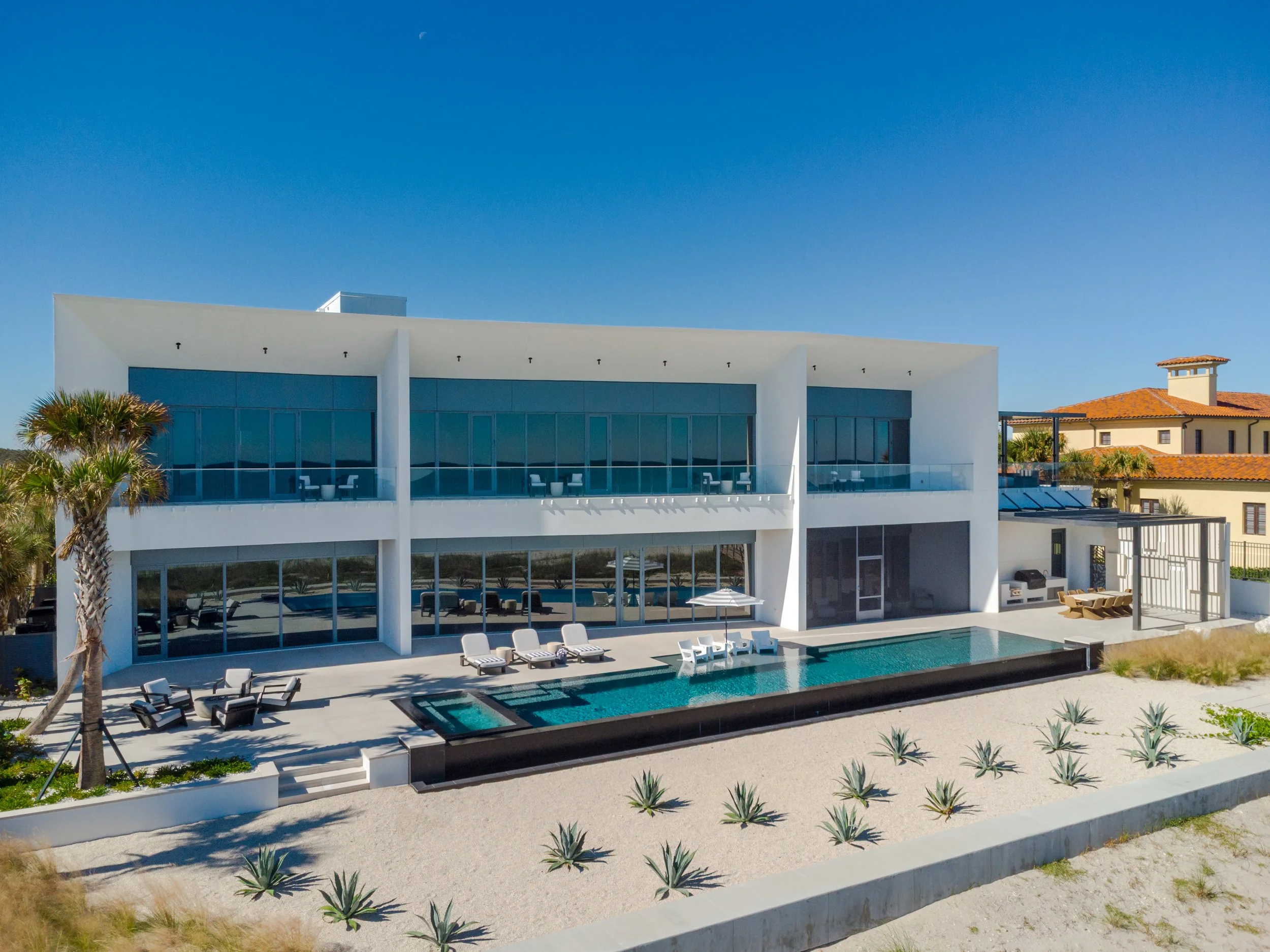 Modern white building with large glass windows and a balcony, outdoor swimming pool with lounge chairs, palm trees, and a sandy area with plants, under a clear blue sky with a visible moon.