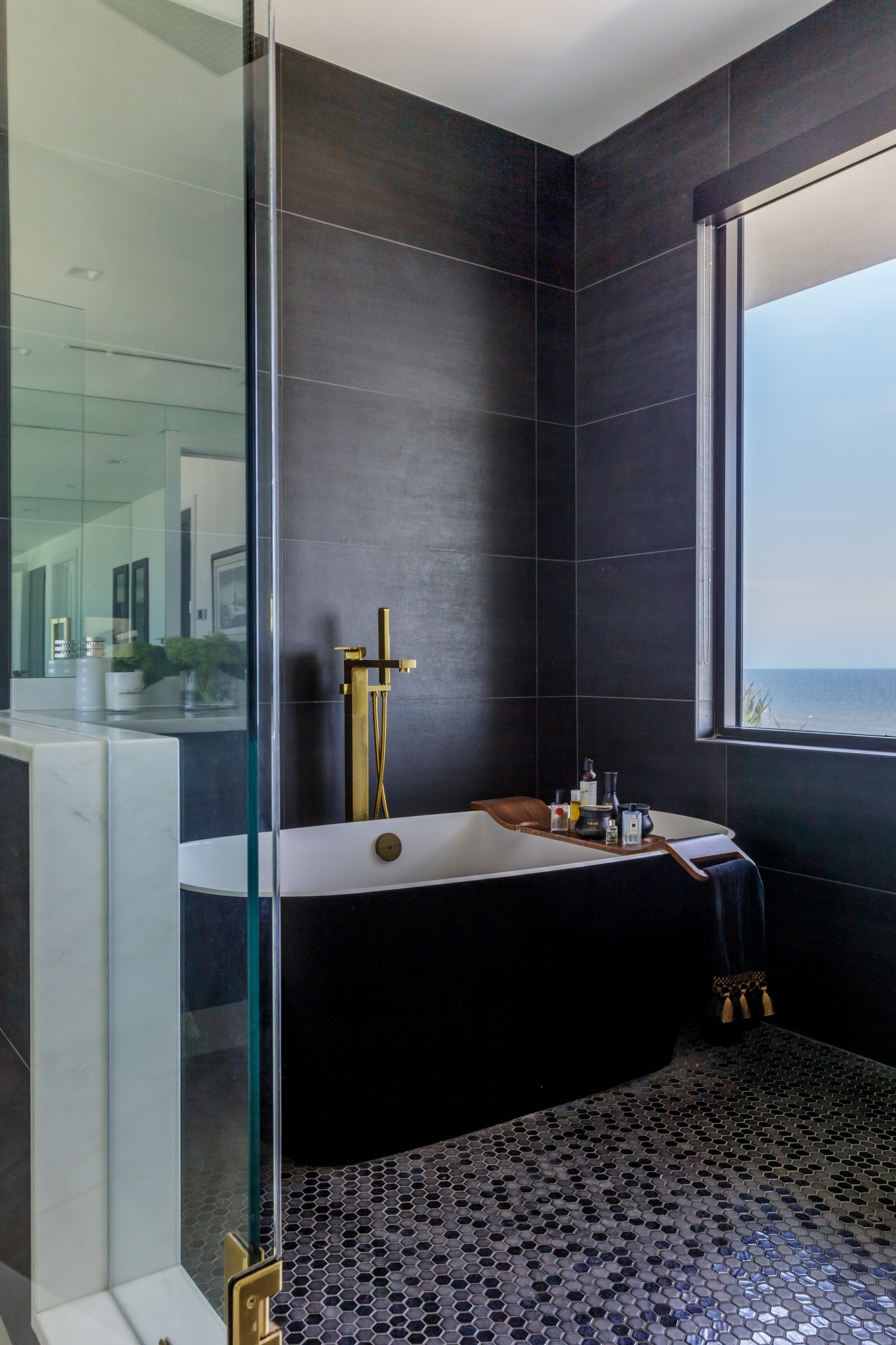 Modern bathroom with black tiled walls and a black soaking tub near a large window with ocean view. A gold faucet and a tray with toiletries are on the tub. A glass shower door is on the left. There's a black hexagonal tile floor.