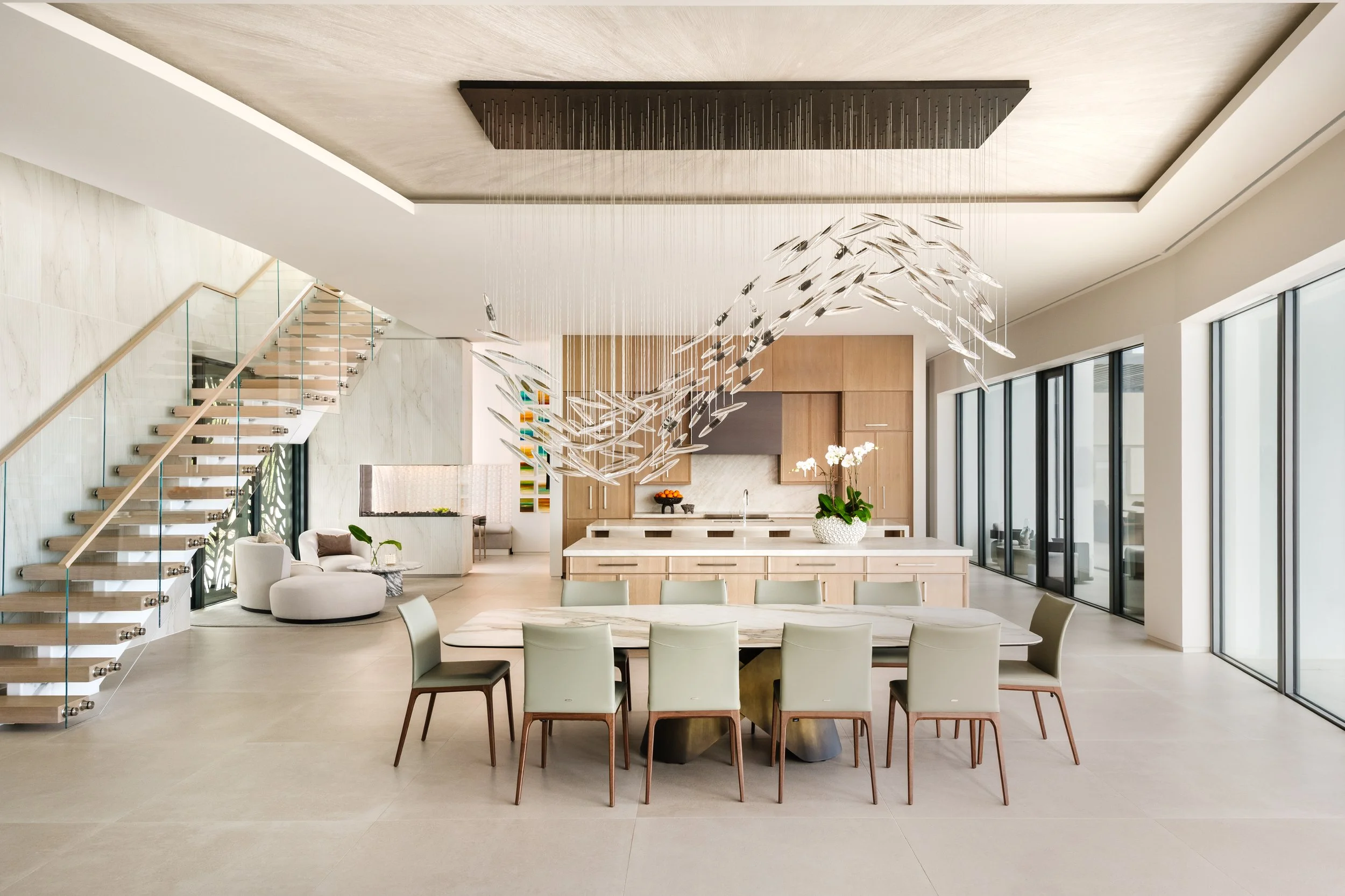 Open-concept modern kitchen and dining area with a decorative chandelier, white chairs around a marble table, and a staircase with glass railing