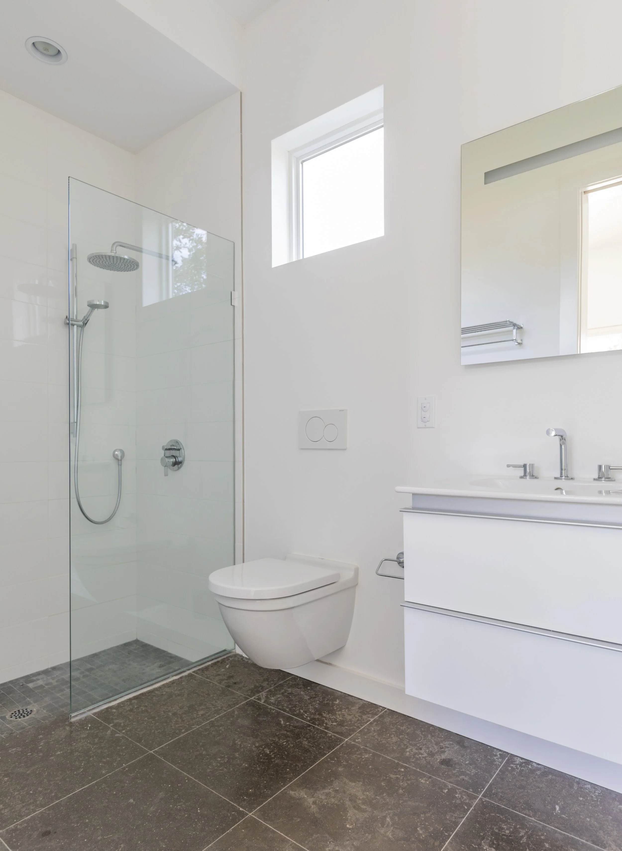 Modern bathroom with walk-in shower, white toilet, white vanity with sink, large mirror, small square window, and dark brown tiled floor.