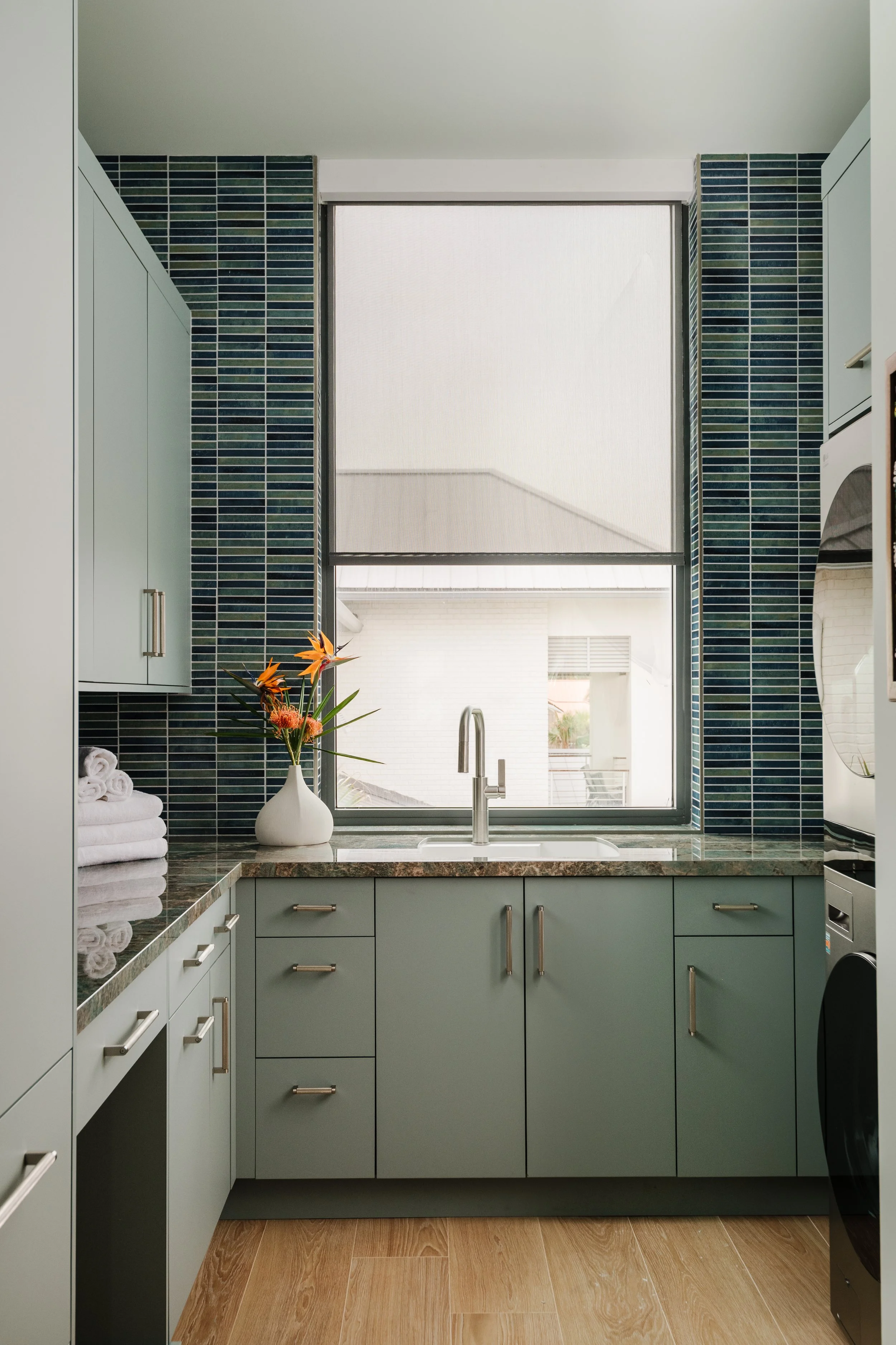 Kitchen with green cabinets, a window with a roller shade, a vase of flowers, towels, and a granite countertop.