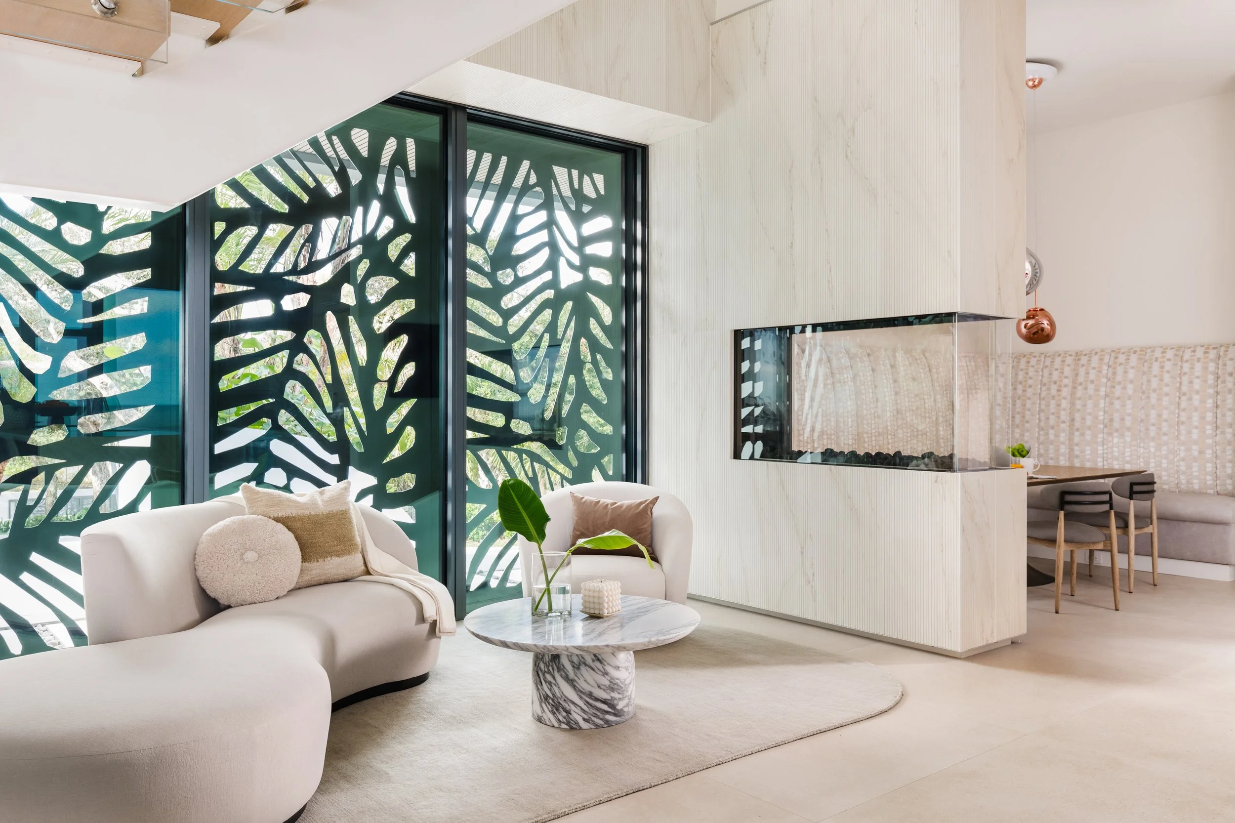Modern living room with white sofa, circular marble coffee table, decorative pillows, green plants, and a black metal room divider with a leaf pattern. Large windows let in natural light.