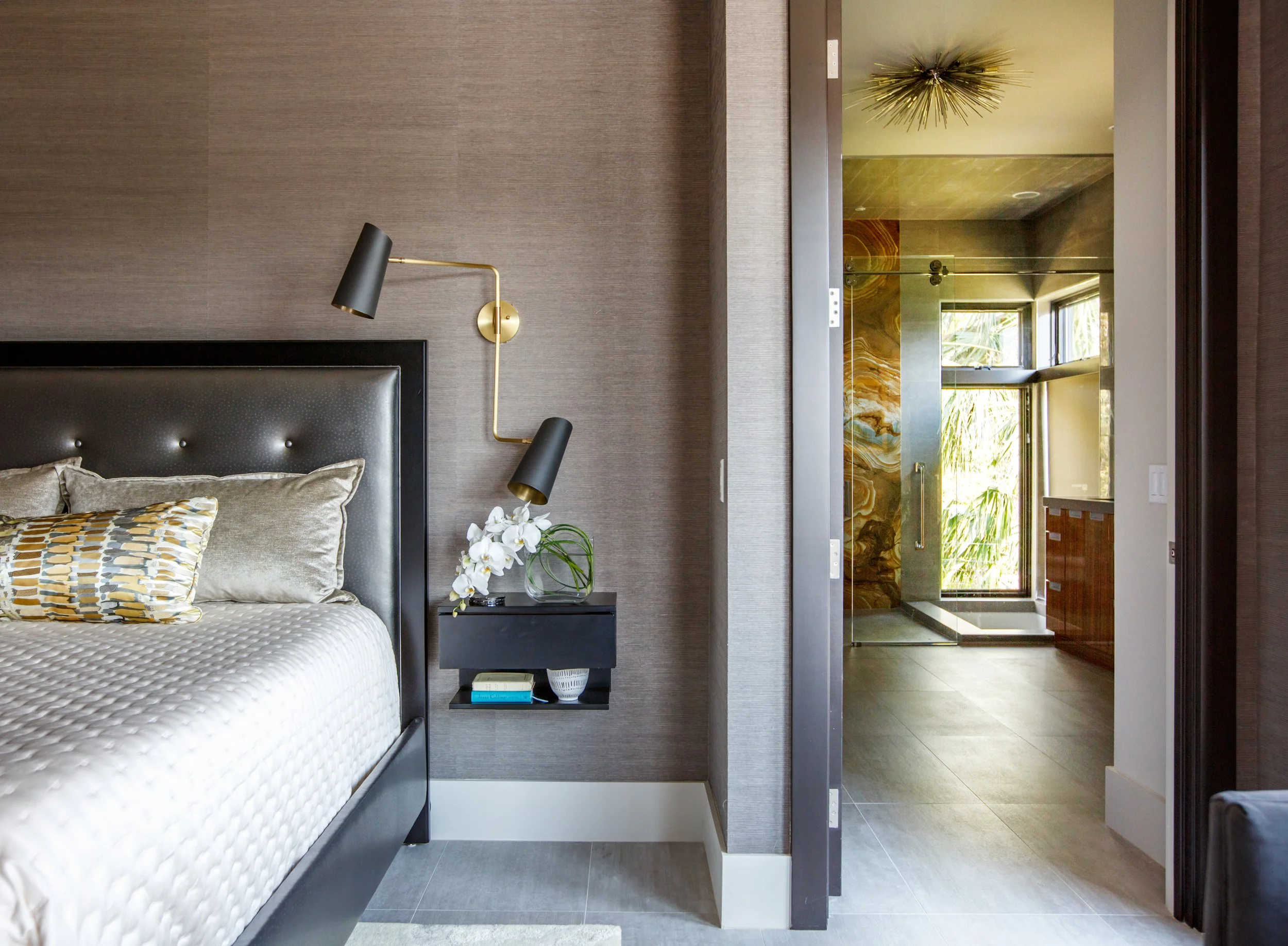 Modern bedroom with a padded headboard, a black bedside table with books and a white orchid, gold wall sconces, and a view into a bathroom with marble walls and large windows.
