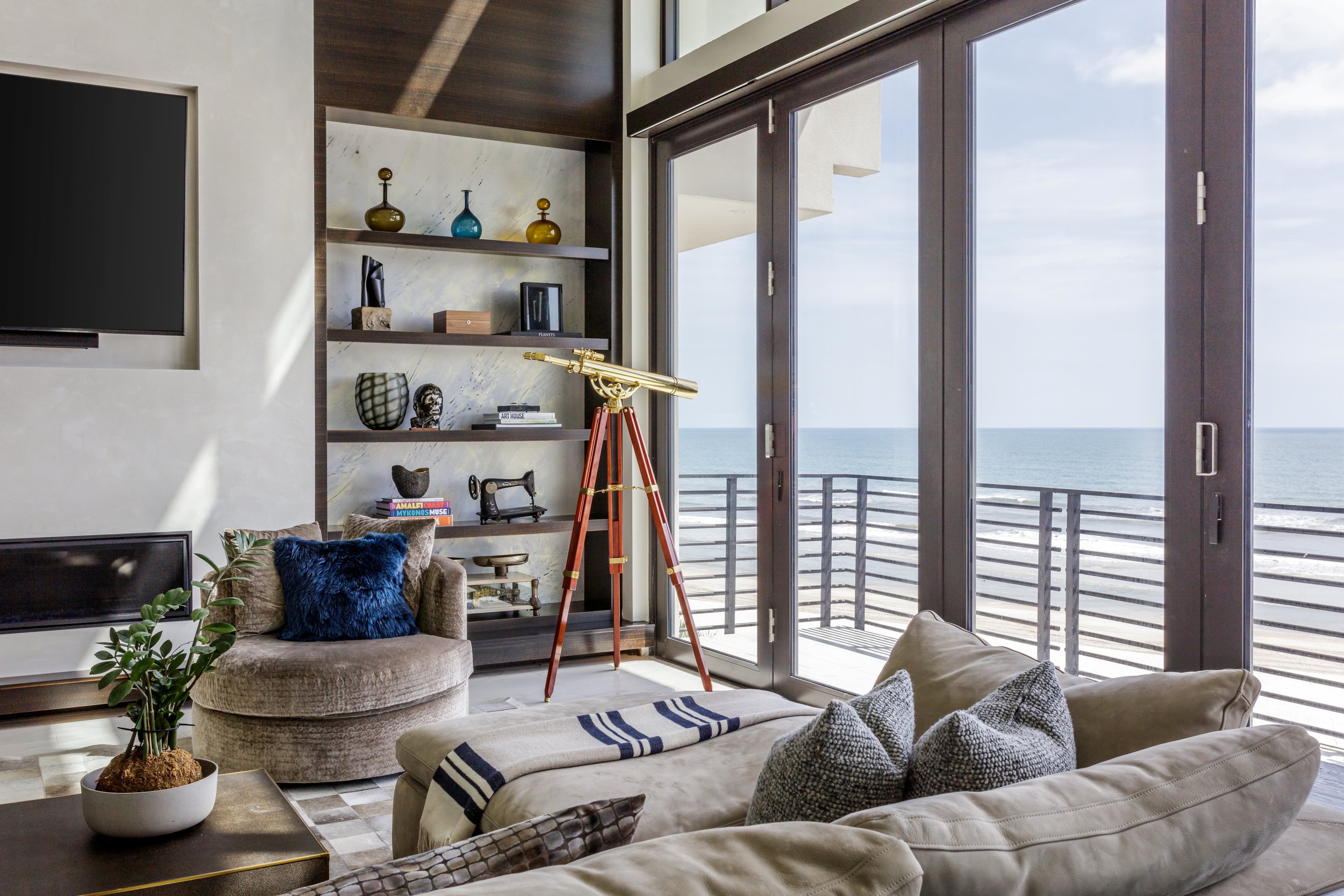Modern living room with large floor-to-ceiling windows overlooking the ocean, decorated with a telescope, books, sculptures, and comfortable seating.