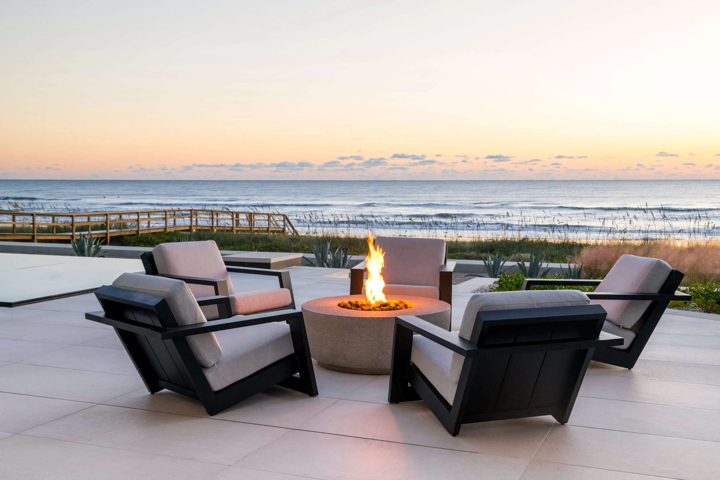 Outdoor patio area with modern chairs surrounding a fire pit, overlooking the ocean at sunset.