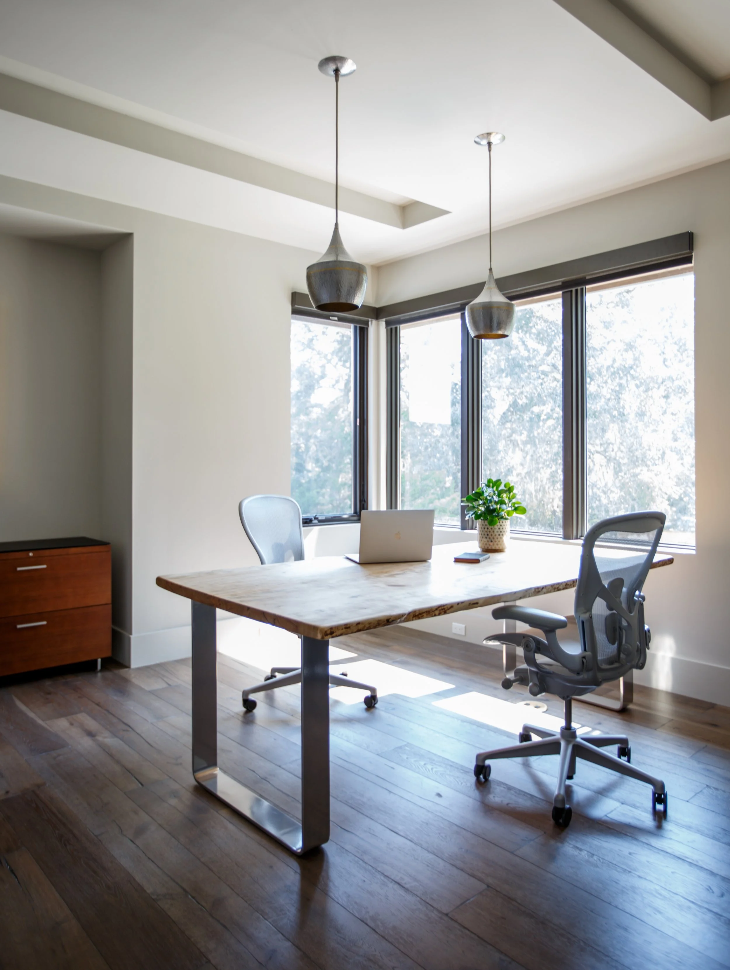 A modern office with a wooden desk, an ergonomic office chair, and large windows allowing natural light, with two pendant lights hanging from the ceiling.