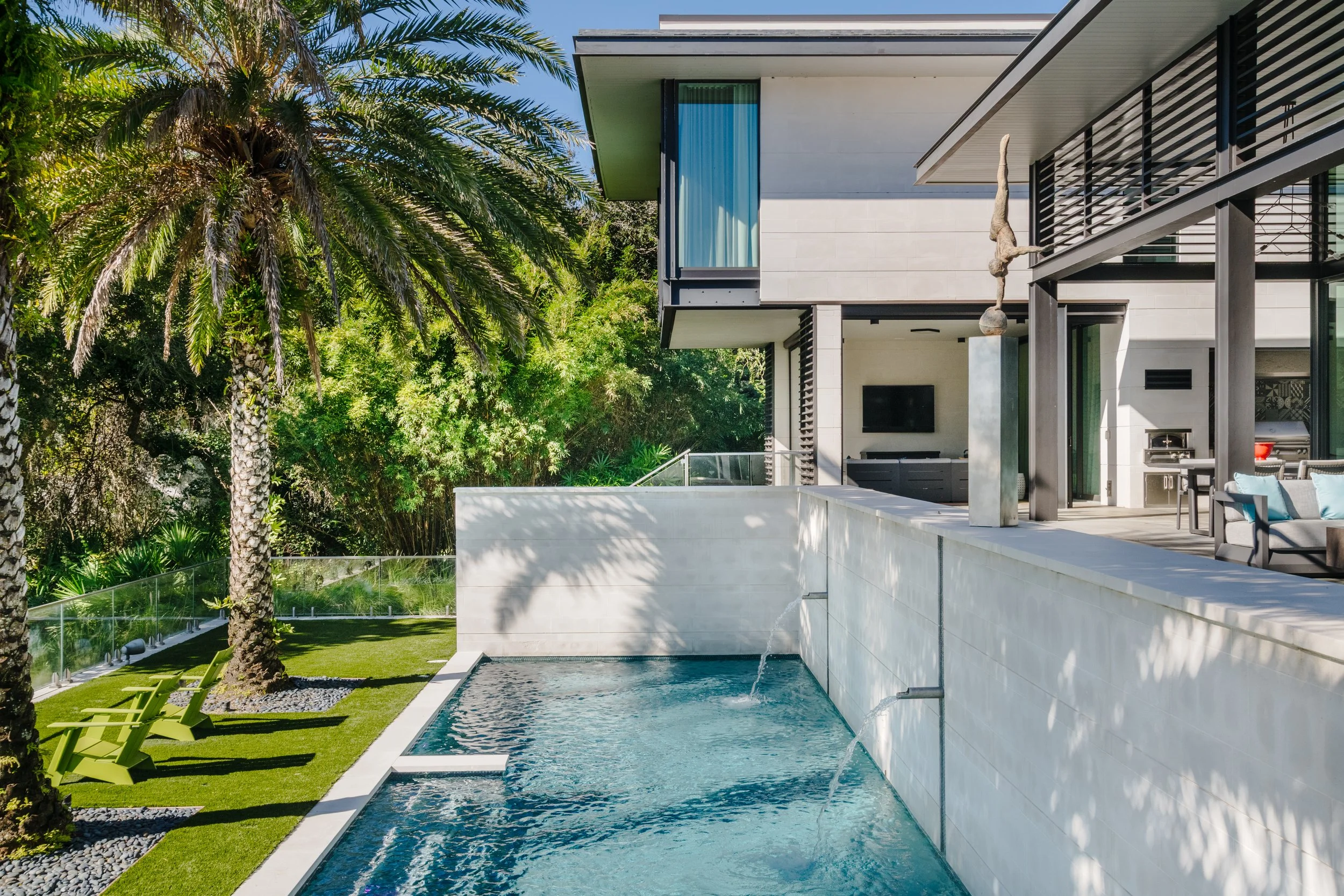 Modern house with white exterior walls and large balconies, surrounded by green trees, with a swimming pool and outdoor lounge area in the foreground.