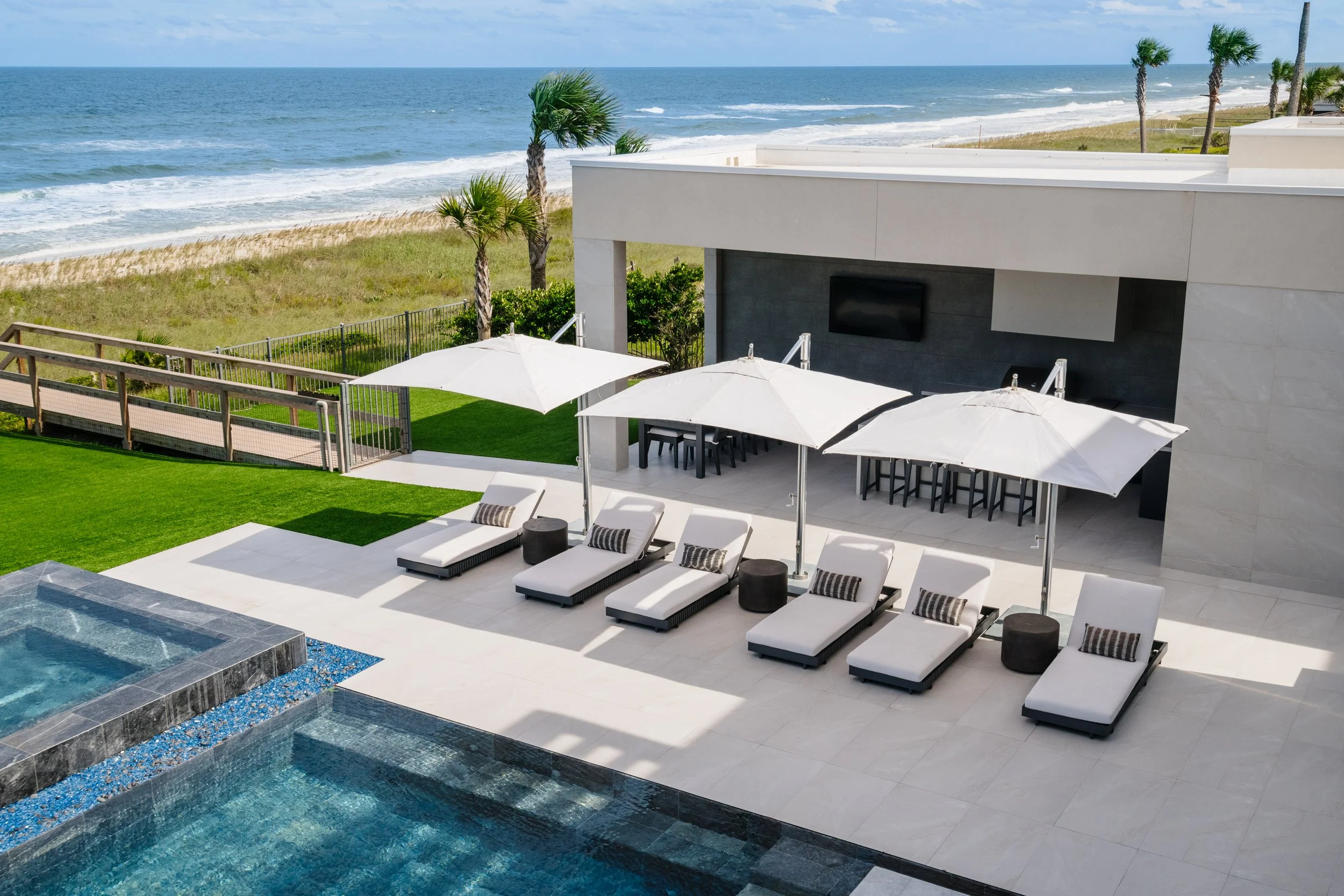A modern beachfront balcony with white lounge chairs and umbrellas, overlooking a sandy beach with palm trees and the ocean in the background.