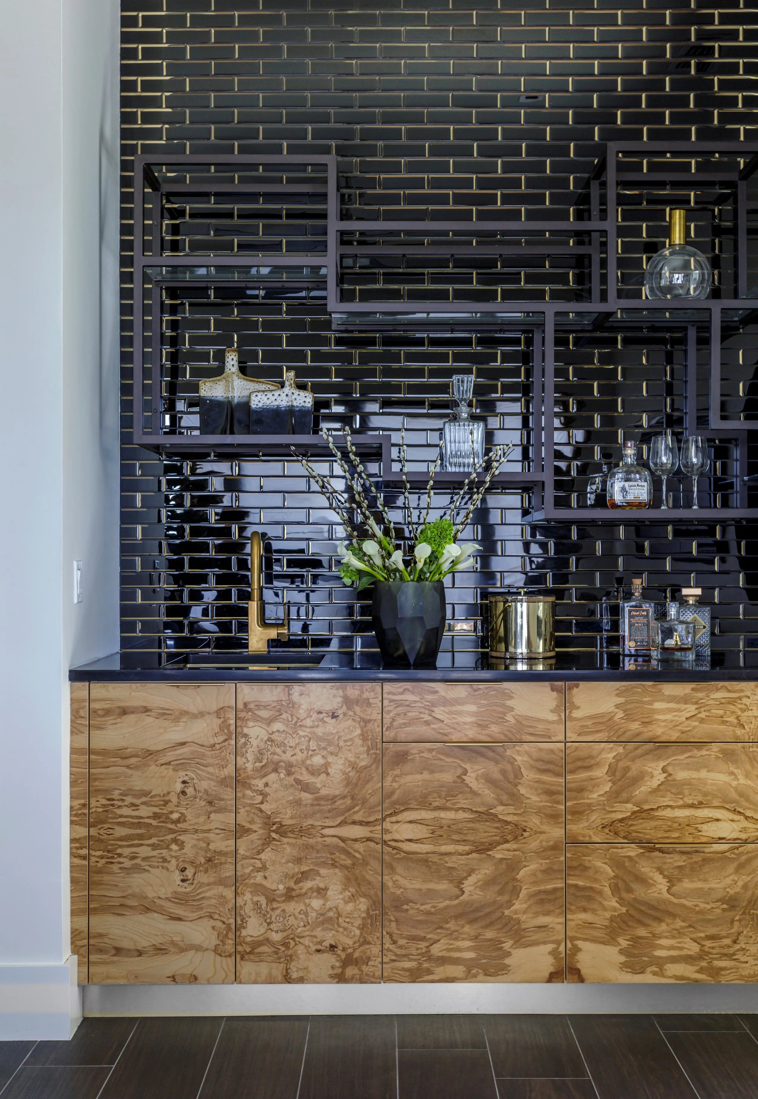 A modern kitchen display with a black tiled wall, light wood cabinet, and decorative items including a black vase with white flowers, bottles, glasses, and a gold-colored faucet.