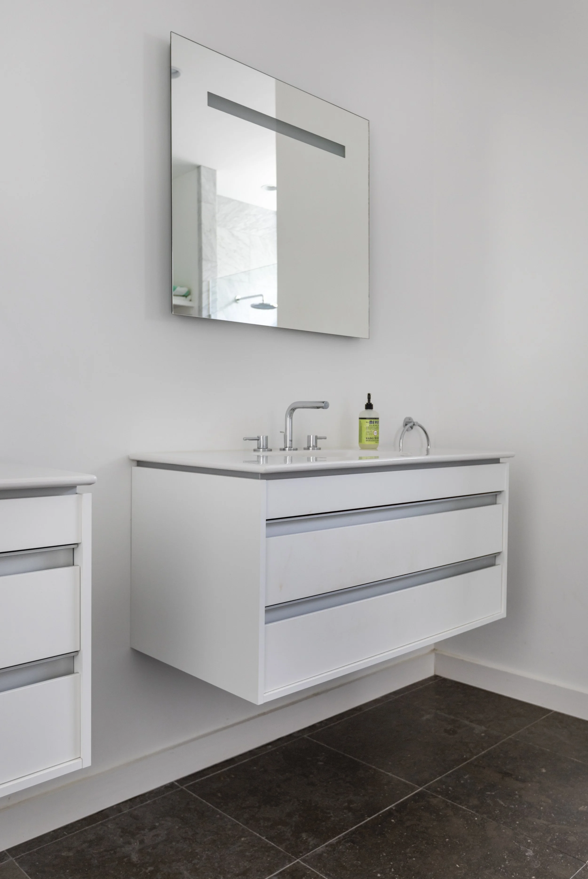 A modern white bathroom vanity with a large drawer and a countertop sink with a silver faucet. There is a black and green hand soap dispenser on the counter. A large rectangular mirror is mounted above the vanity, reflecting part of the bathroom, inc