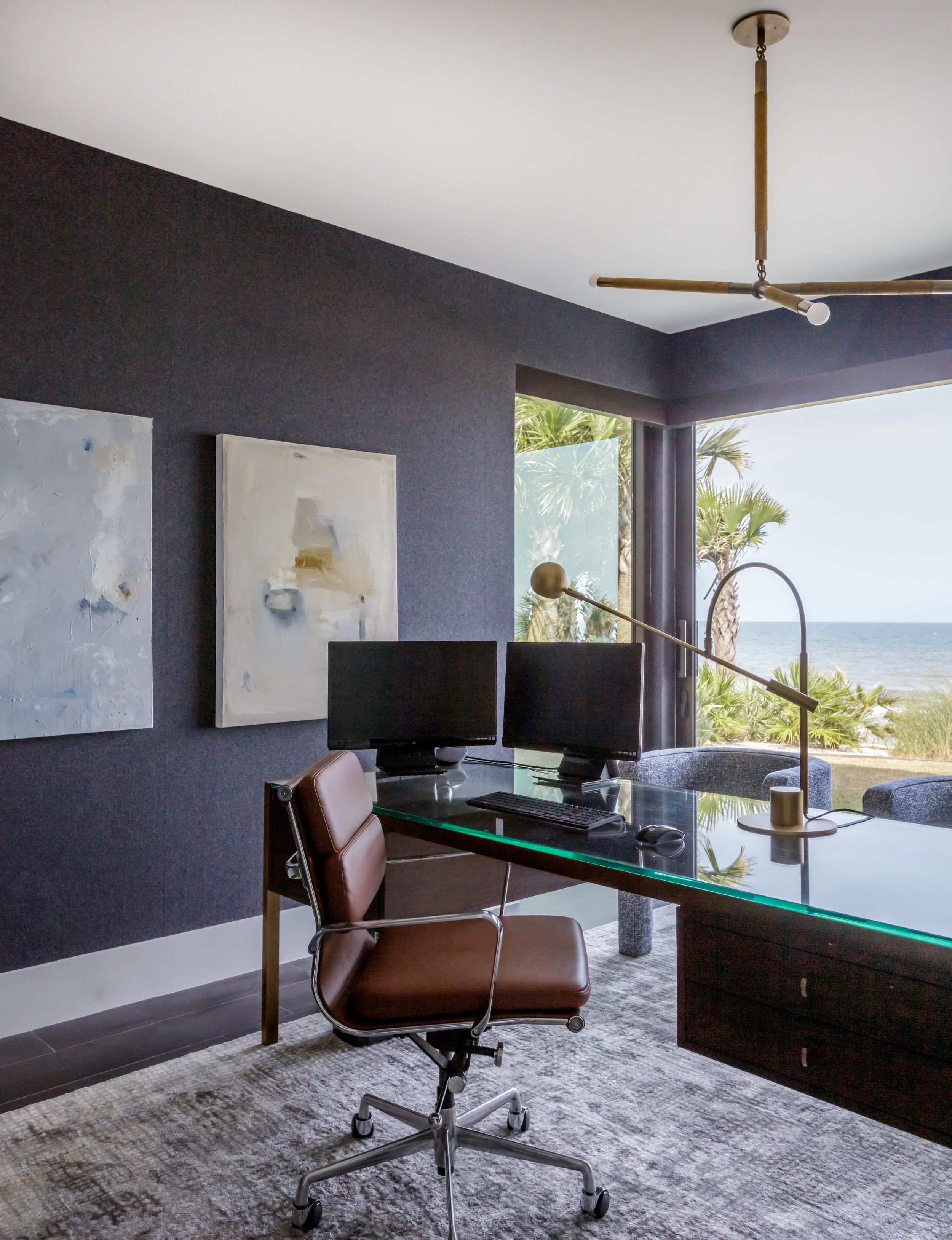 Modern home office with glass desk, brown leather office chair, two computer monitors, white abstract artwork on dark wall, large window overlooking palm trees and ocean, ceiling fan, and lamp, with a patterned area rug.