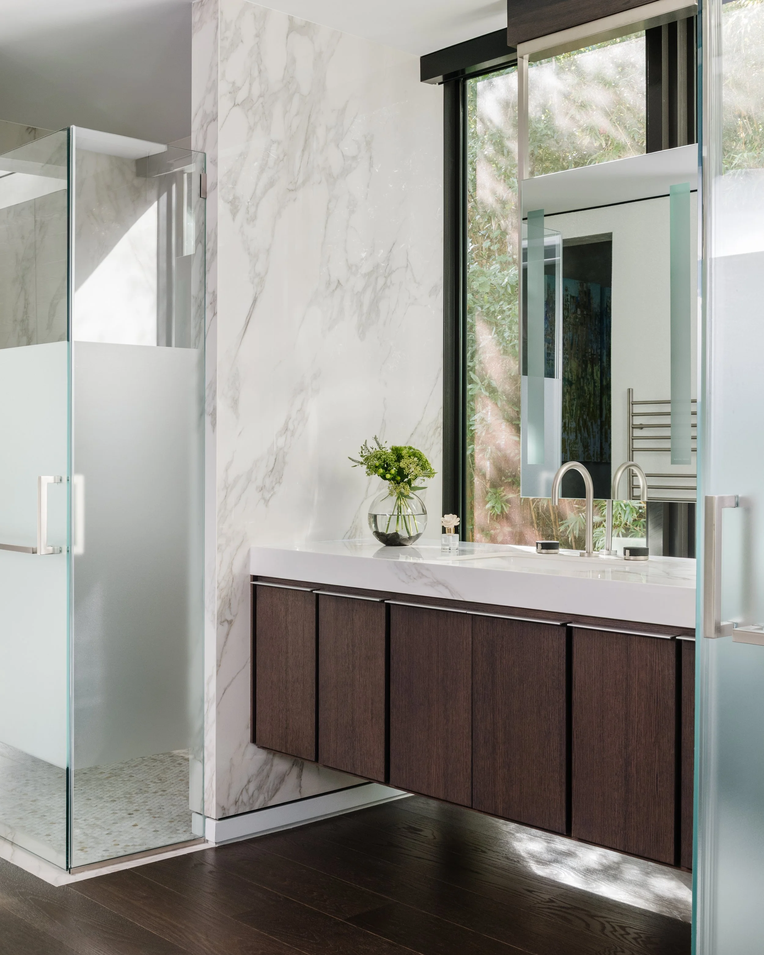 Modern bathroom with a marble wall, glass shower enclosure, dark wood vanity with a marble countertop, a plant in a glass vase, and a large window with greenery outside.