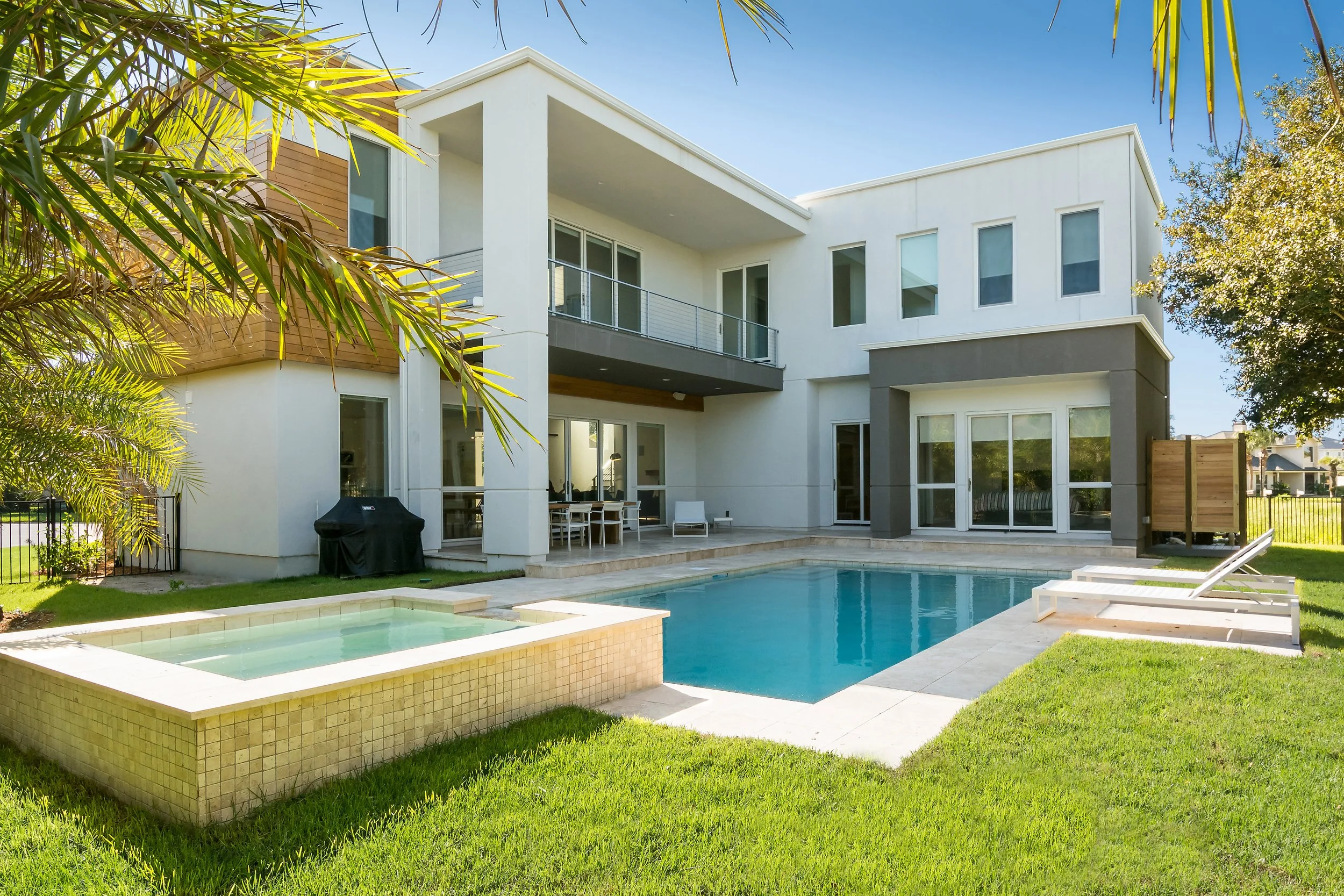 Modern two-story house with large windows, balcony, and backyard with pool, hot tub, lawn, and trees under a clear blue sky.