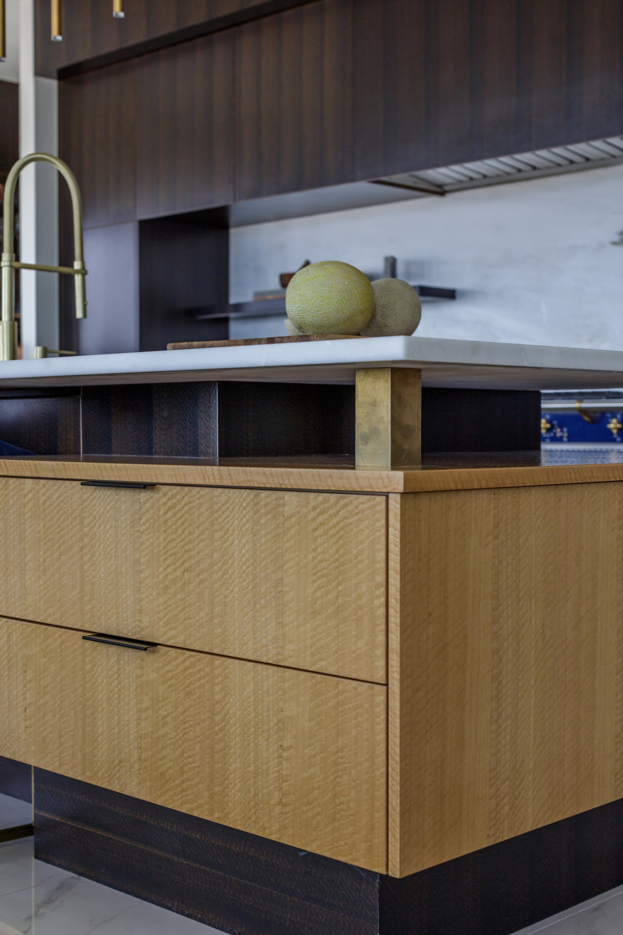 A kitchen counter with a marble surface holding three melons and a wooden cutting board. The cabinet below is light wood with a dark bottom panel, and there is a gold faucet near the counter.
