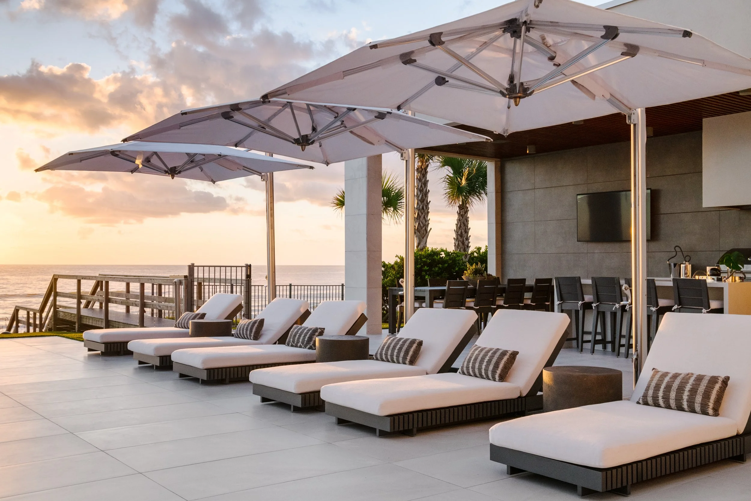 Outdoor lounge area with white sun loungers, striped pillows, and large white umbrellas at sunset, overlooking a beach with palm trees and a cloudy sky.