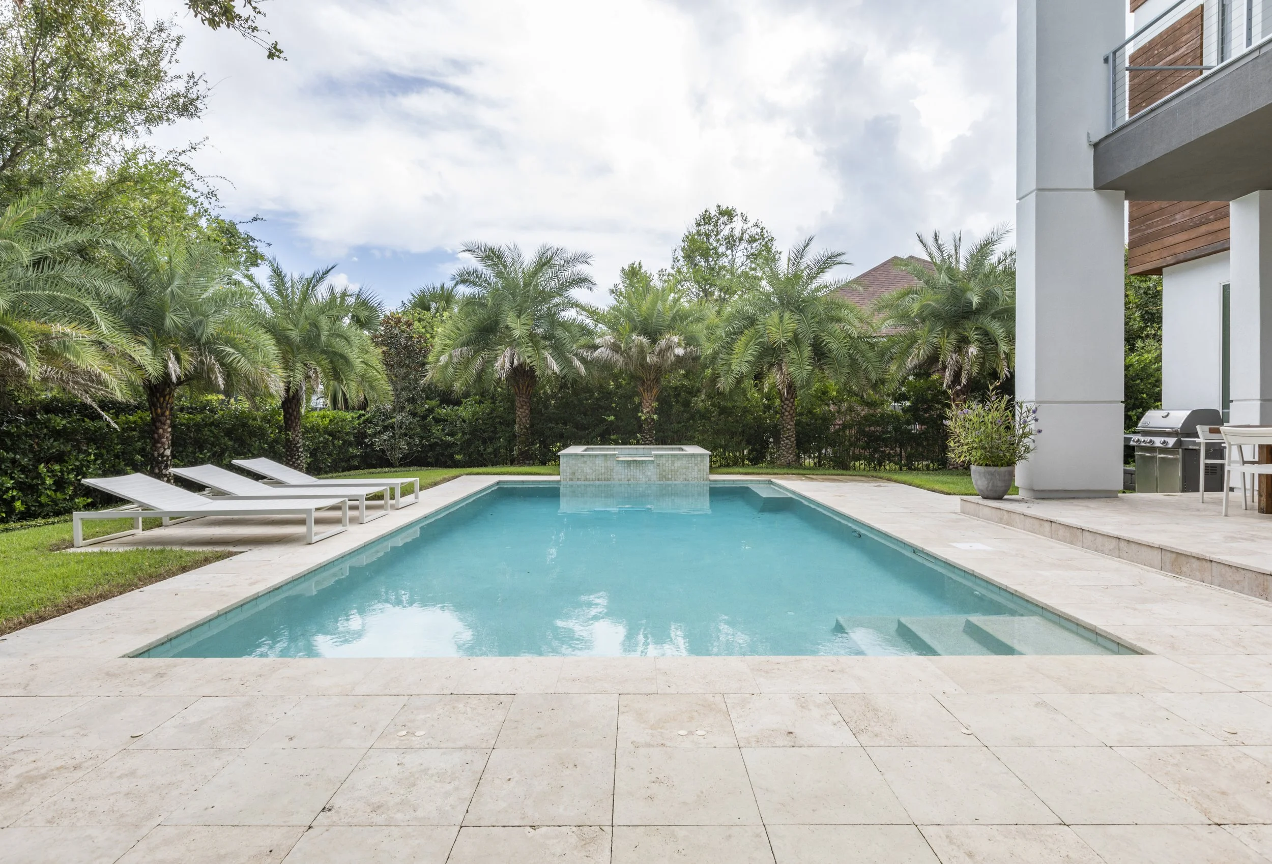 A backyard with a swimming pool, lounge chairs, lush palm trees, and modern residential buildings.