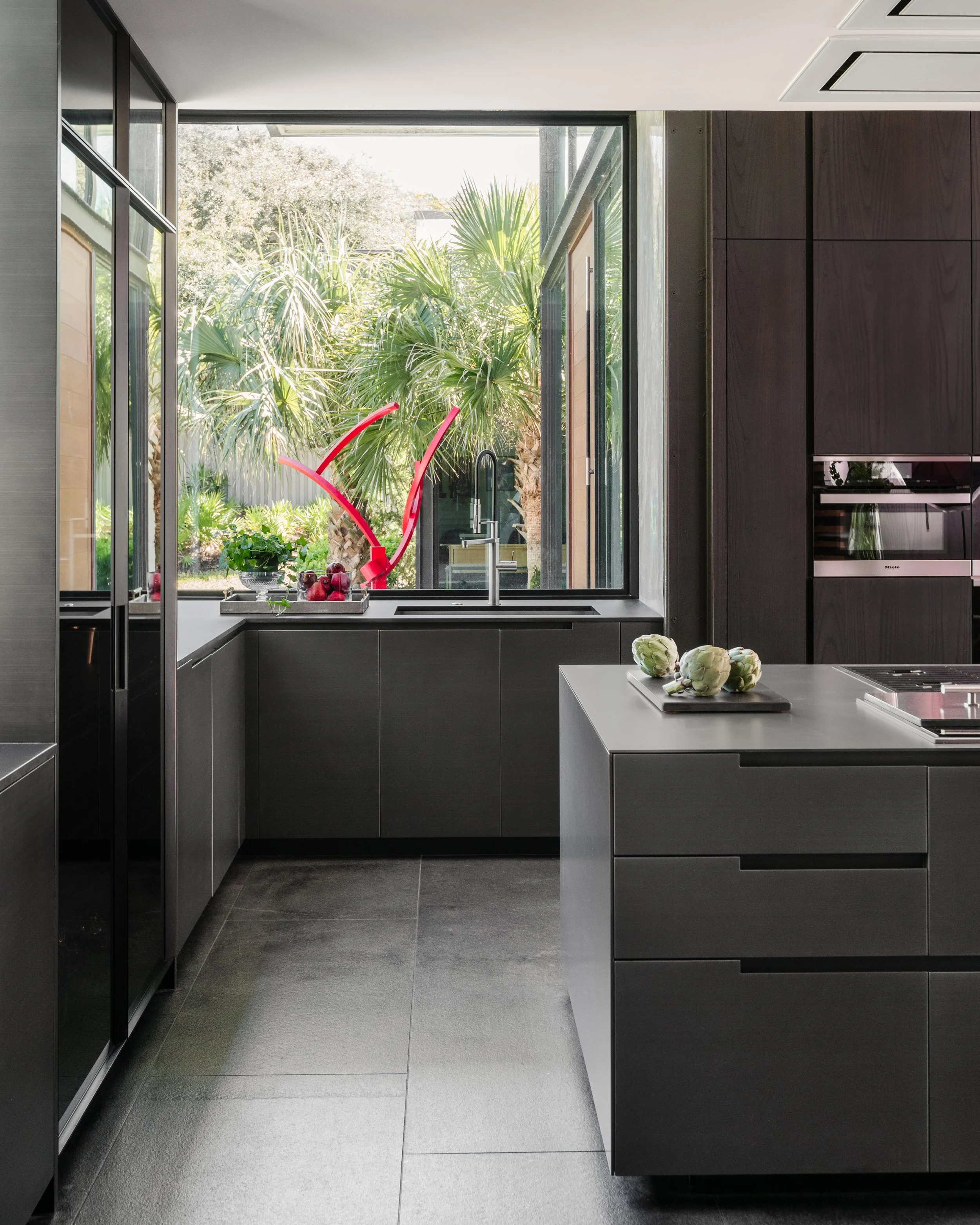 Modern kitchen with dark cabinetry, grey countertops, a large window with a view of outdoor trees, and a table with artichokes and a cutting board.