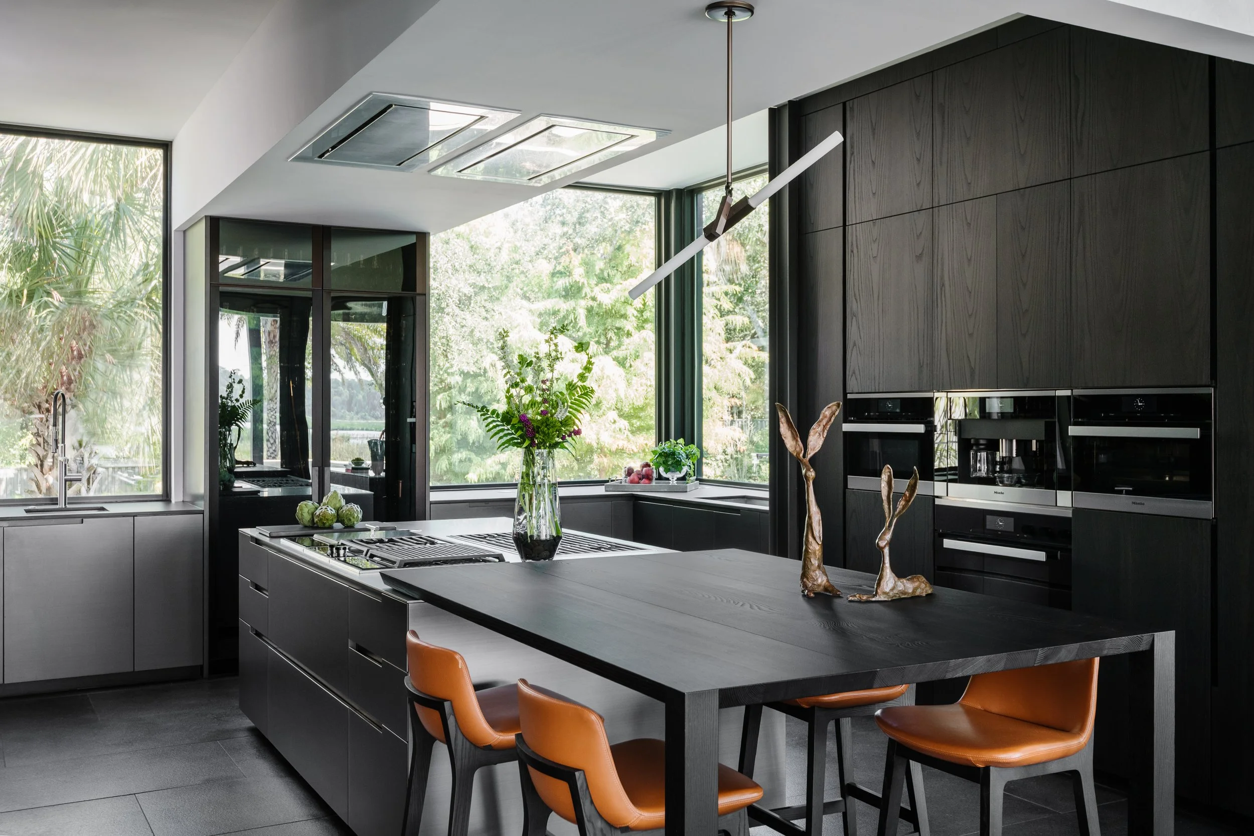 Modern kitchen with dark cabinetry and large windows, decorated with a vase of flowers on the island and orange cushioned chairs around a dining table.
