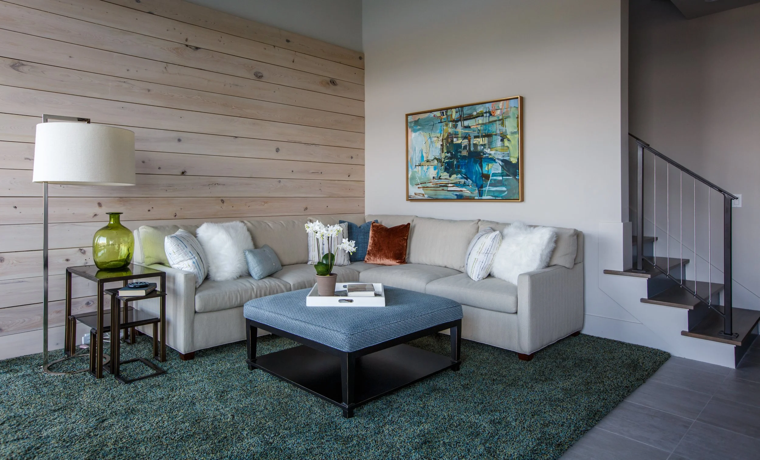 Living room with a beige sectional sofa decorated with pillows, a blue ottoman, a floor lamp with a white shade, a side table with a green vase and a remote, a wall artwork with abstract blue and green shapes, a wooden-paneled wall, and staircase wit