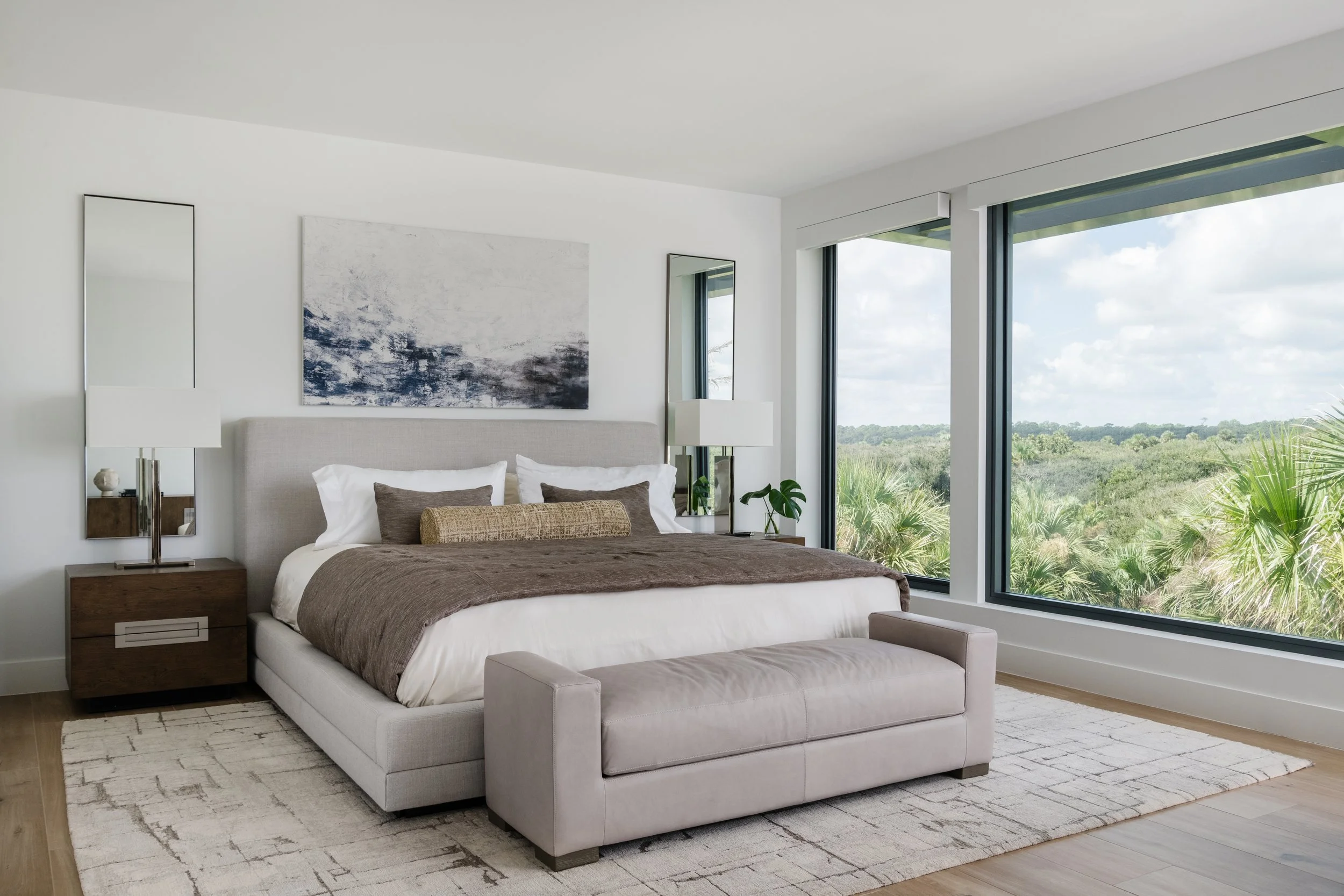 A modern bedroom with a large bed, white bedding, and various pillows, flanked by two nightstands with lamps. Large windows reveal a lush green landscape outside.