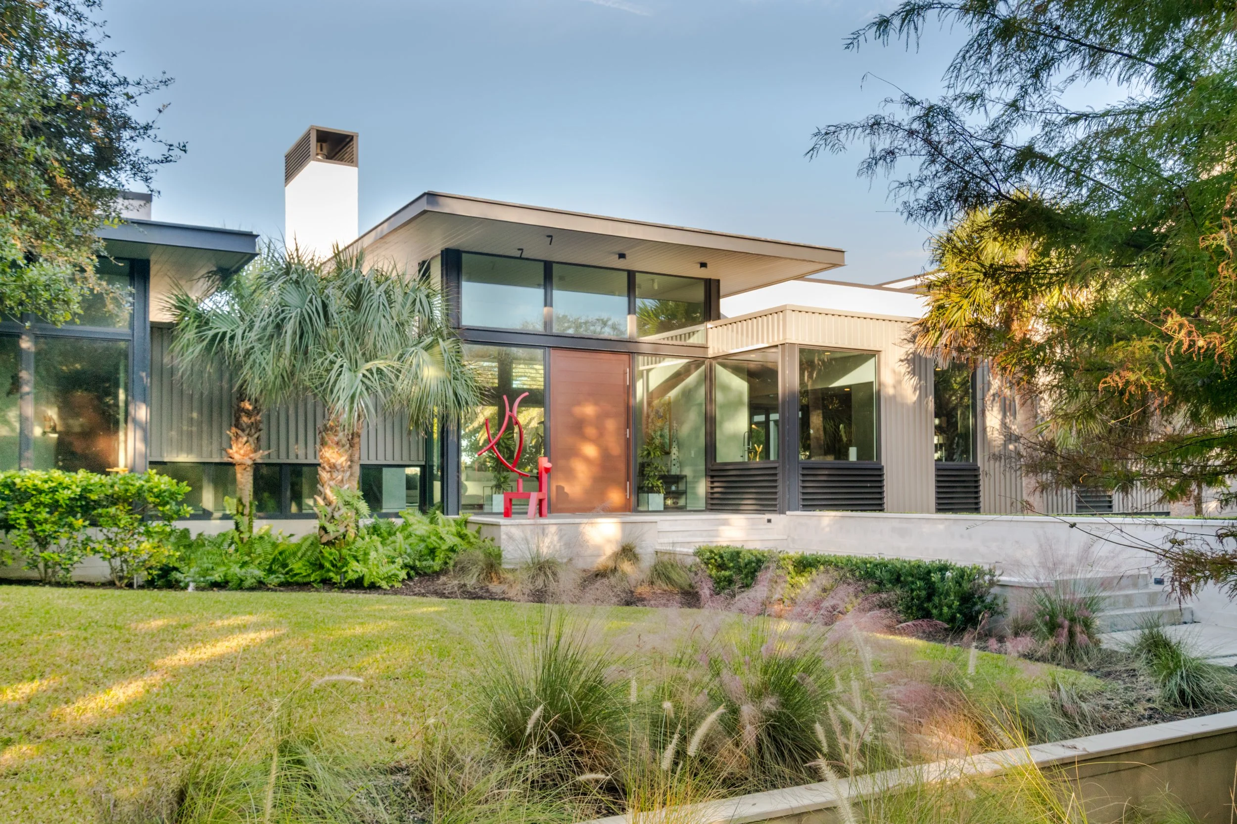 Modern two-story house with large windows, surrounded by a lush garden with various trees and plants, under a clear blue sky.