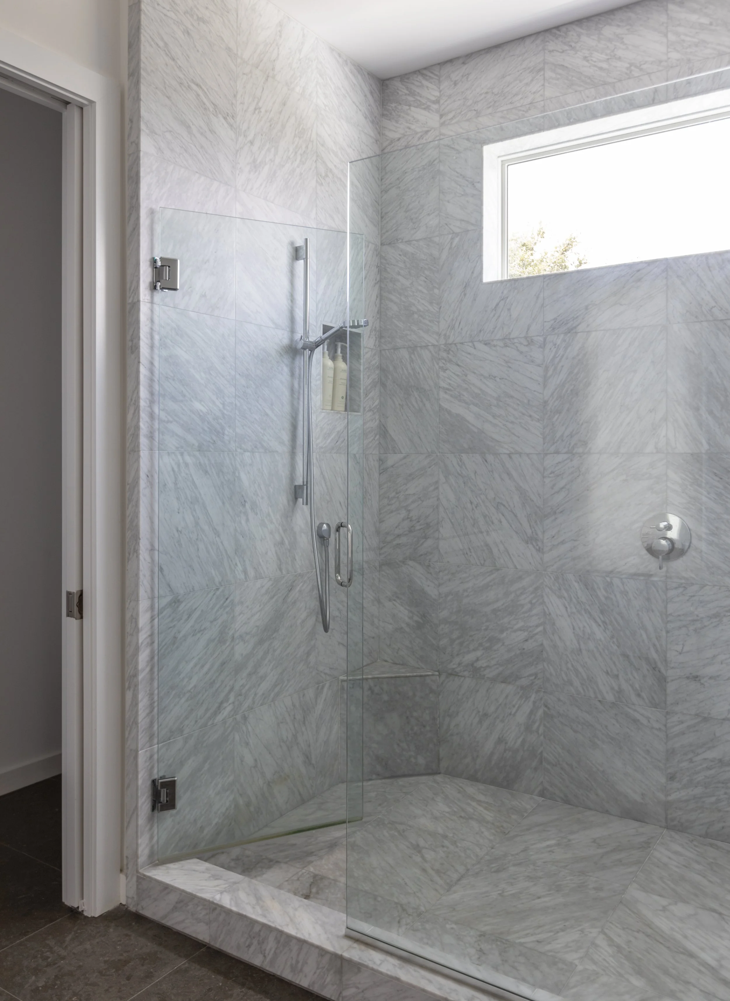 Modern walk-in shower with white marble tiles, glass door, and a high window letting in natural light.