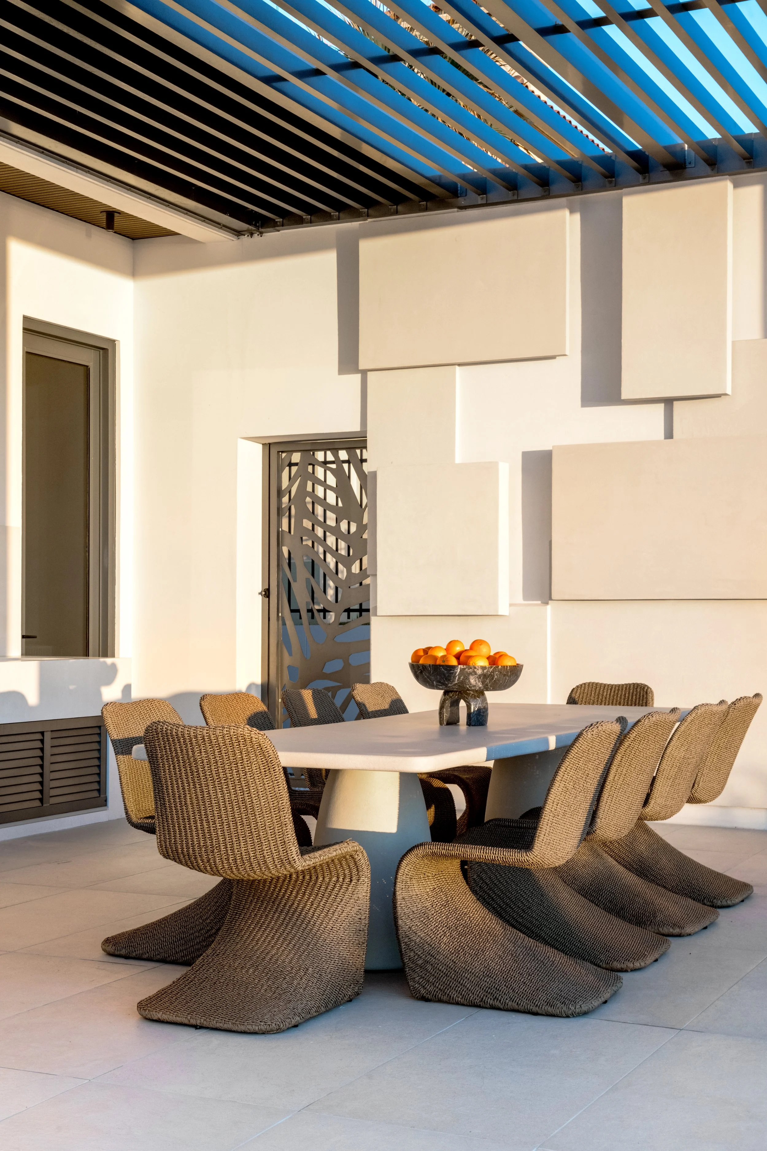 An outdoor patio with a white table surrounded by tan woven lounge chairs. There is a black bowl with oranges on the table, and a wall with geometric decor and a door in the background. Blue and black slatted shading overhead.