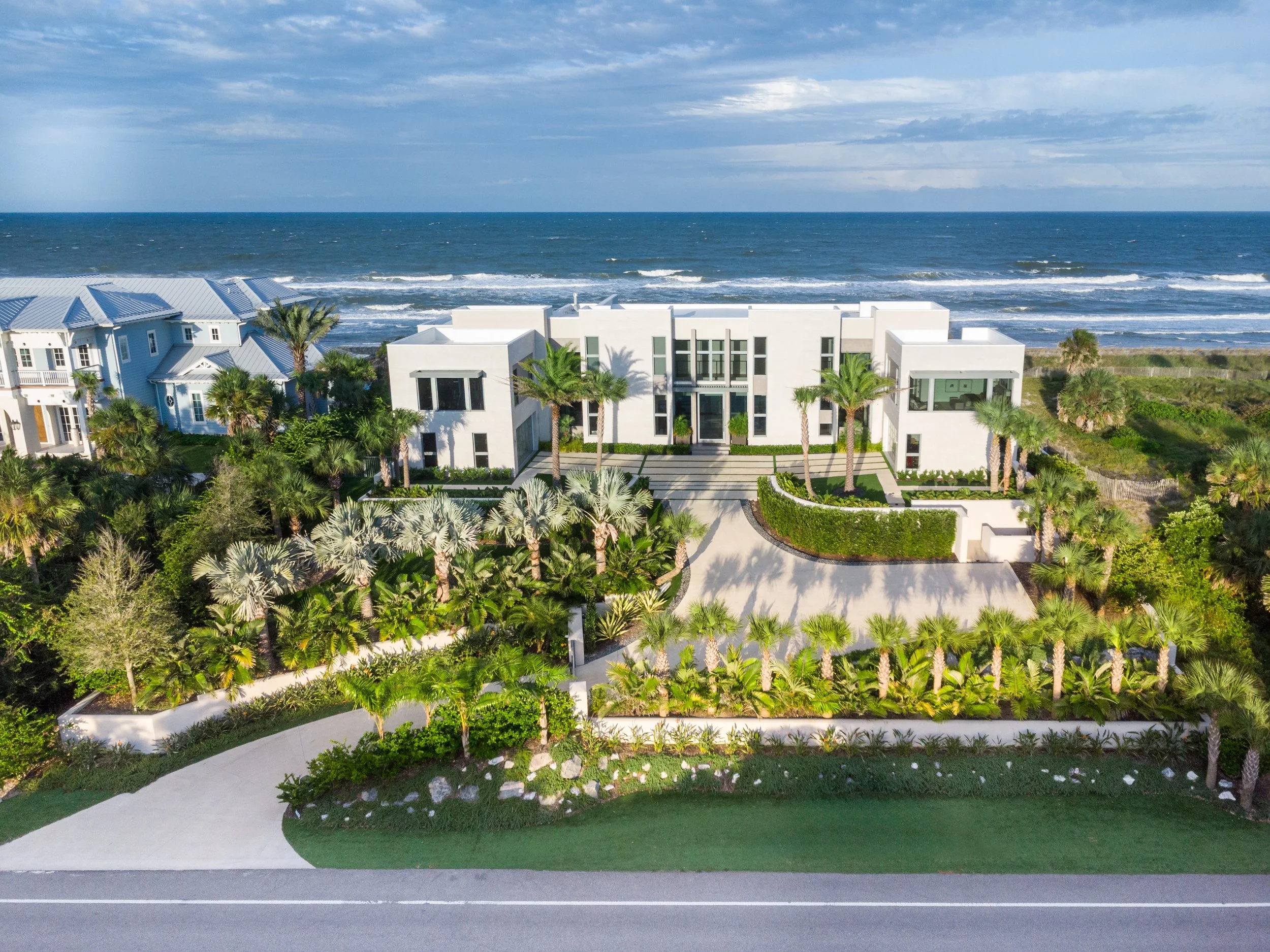 Aerial view of modern white beachfront house with palm trees, landscaped garden, and a road in front, with the ocean and cloudy sky in the background.