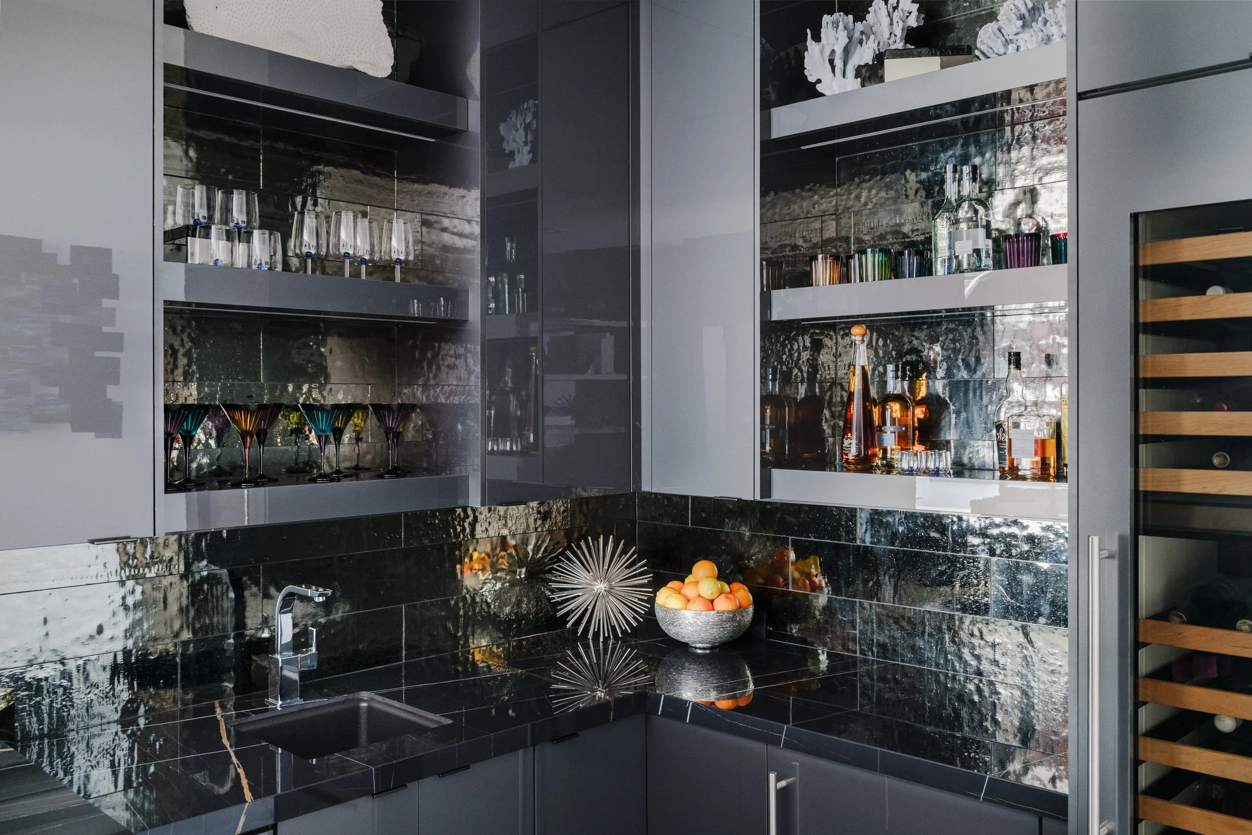 Modern kitchen with black marble countertops and backsplash, glassware and liquor bottles on shelves, bowl of oranges, and decorative items.