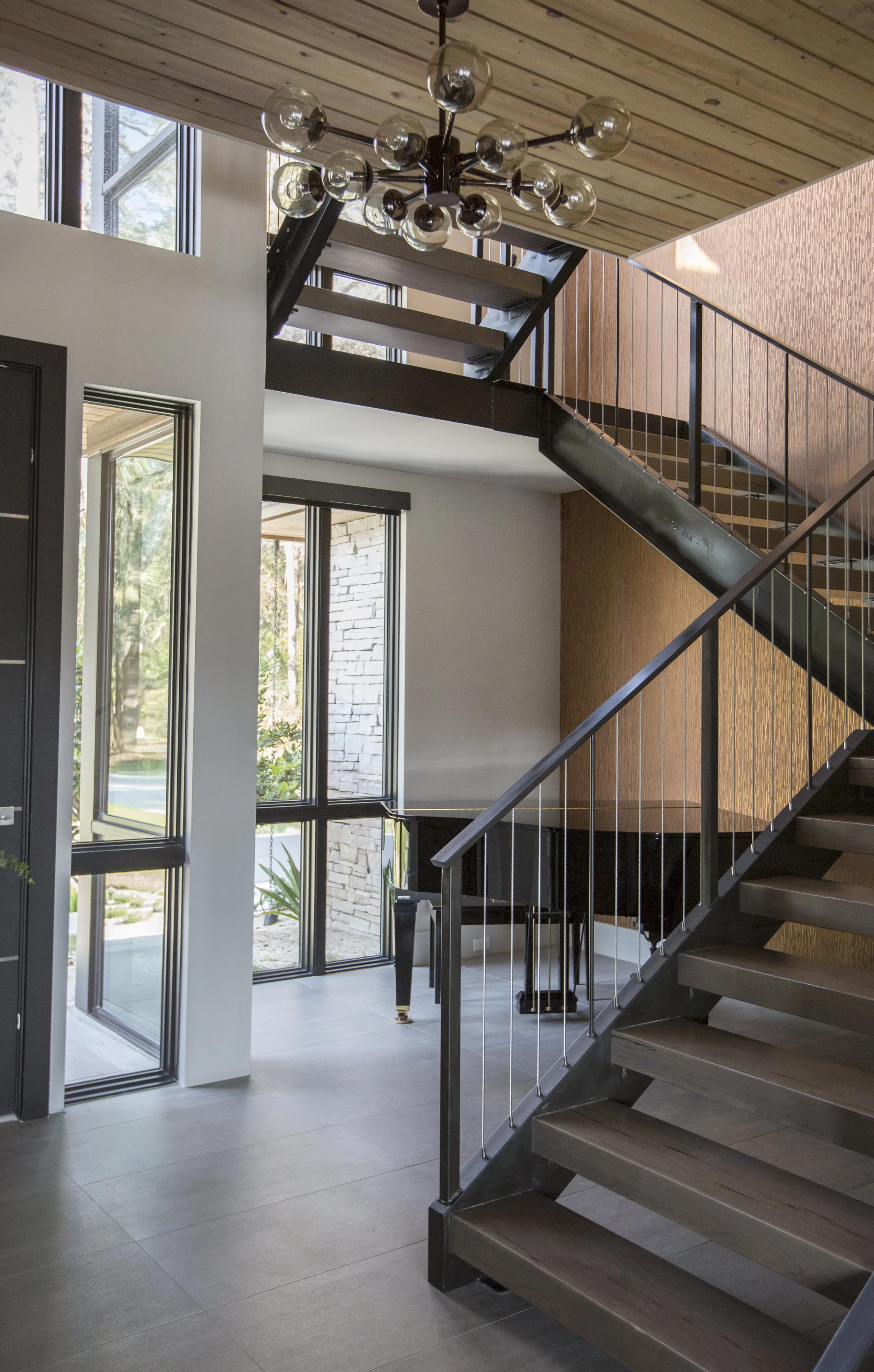Interior view of a modern home with a staircase, large windows, and a wood-paneled ceiling, with a contemporary chandelier hanging from the ceiling.