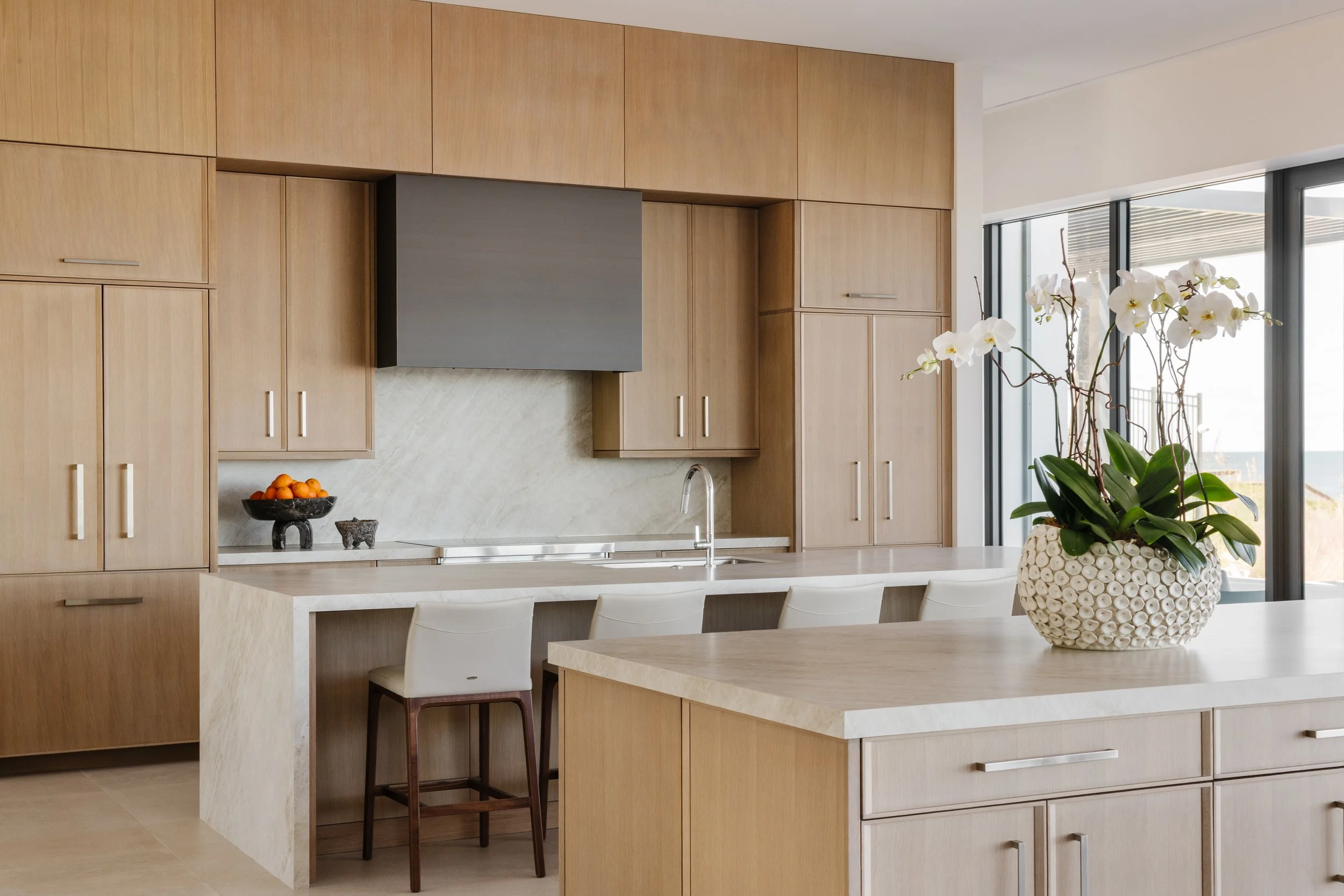 Modern kitchen with wooden cabinets, white marble countertops, a bowl of oranges, and a white orchid plant in a textured vase. Large windows provide natural light.