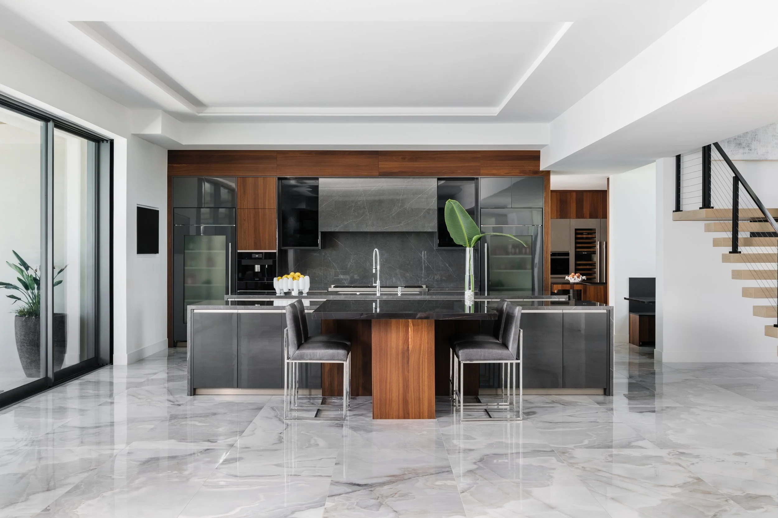 Modern kitchen with wooden and black cabinets, marble backsplash, marble floor, black dining table with black chairs, large sliding glass door, stairway to the right