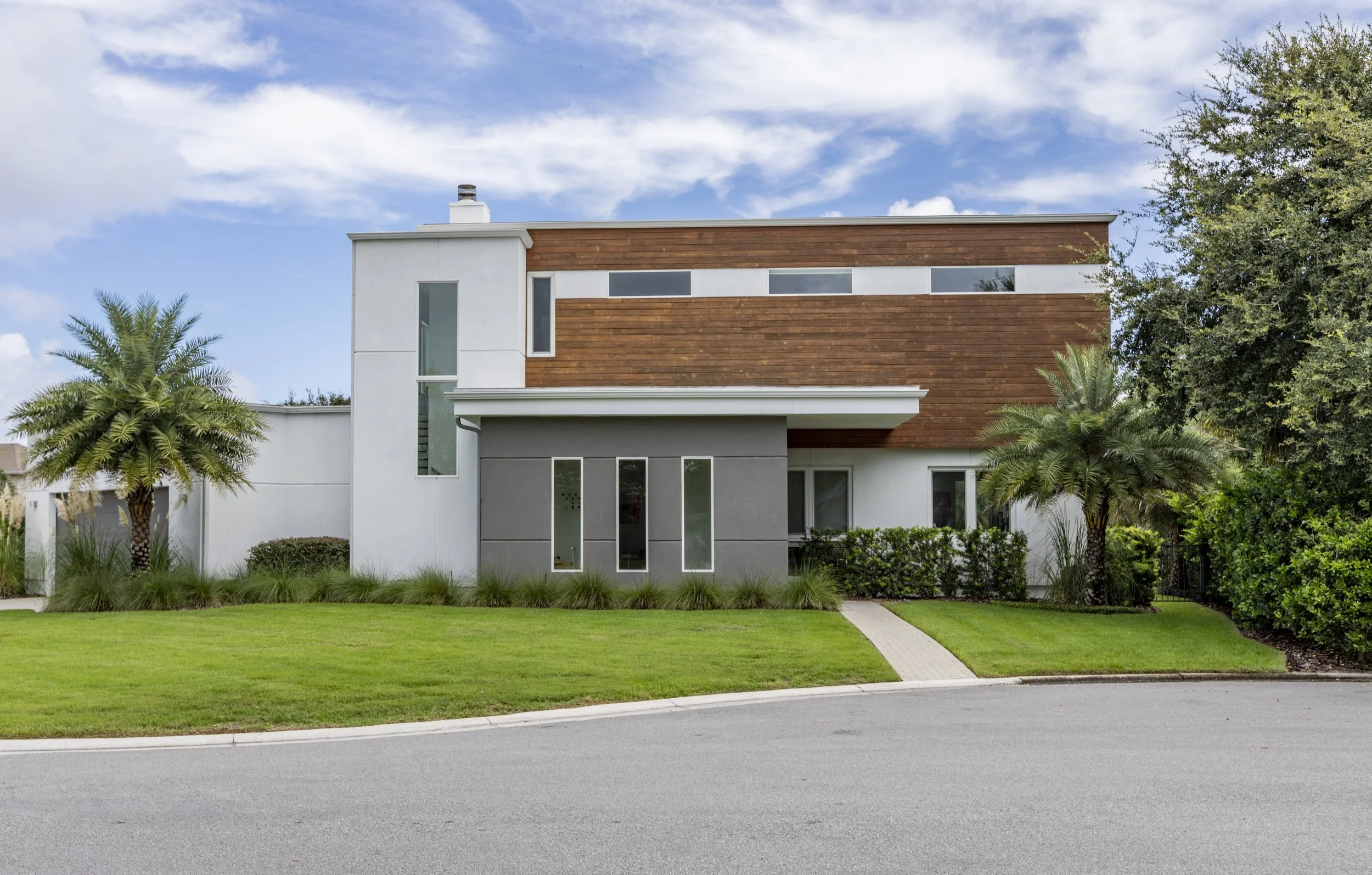 Modern residential house with white and gray exterior, wooden accents, and large windows, surrounded by green grass, palm trees, and other lush vegetation, under a partly cloudy sky.