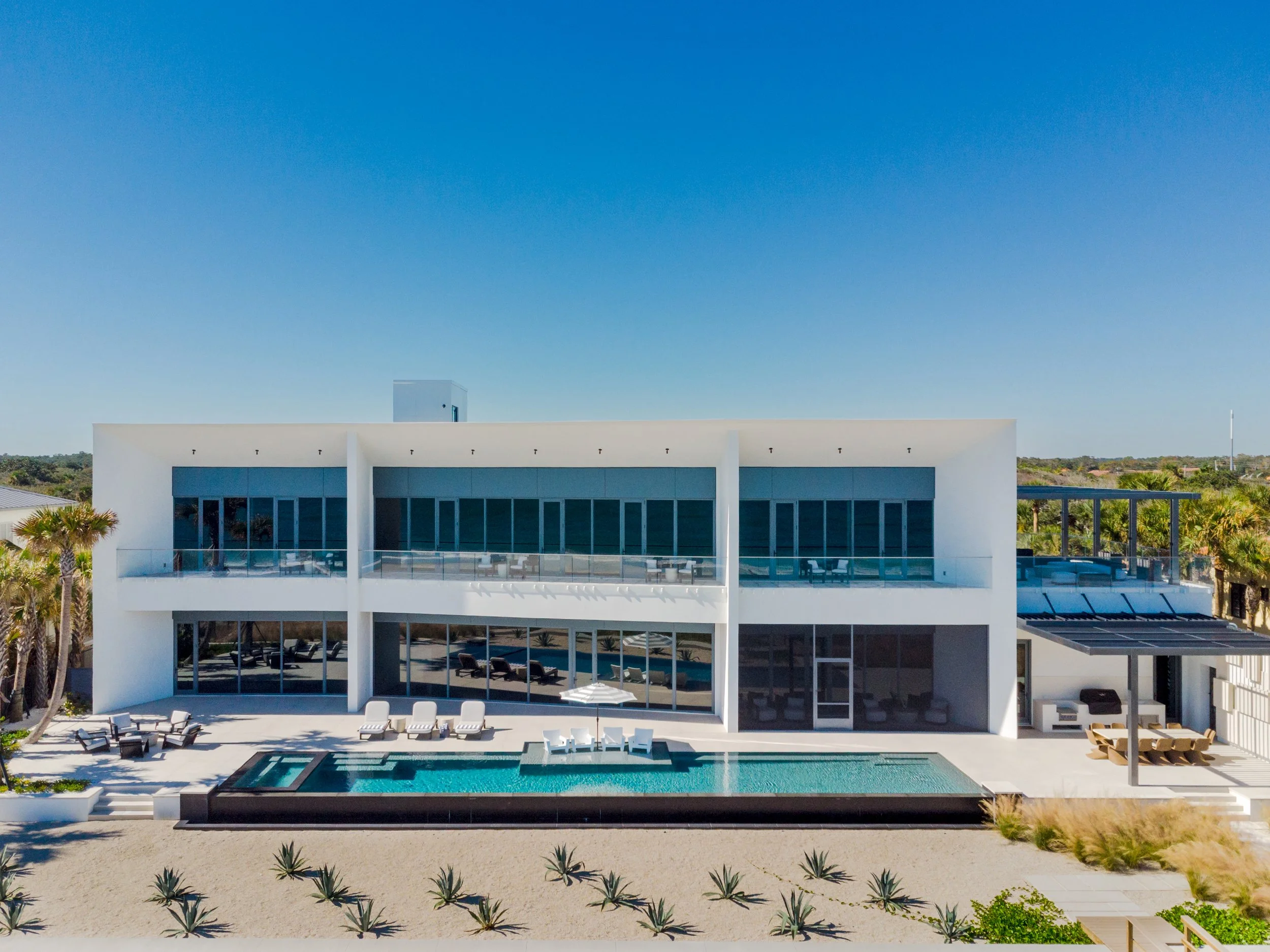 Modern white house with large glass windows, outdoor pool, and patio furniture, set against a clear blue sky with surrounding palm trees.