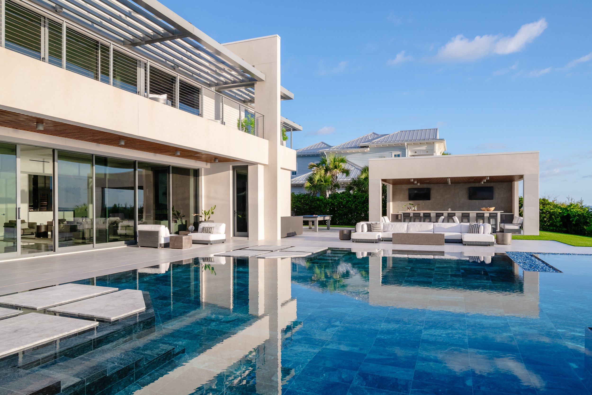 Modern house with large outdoor pool, outdoor seating area, and balcony with metal railings, under a clear blue sky.