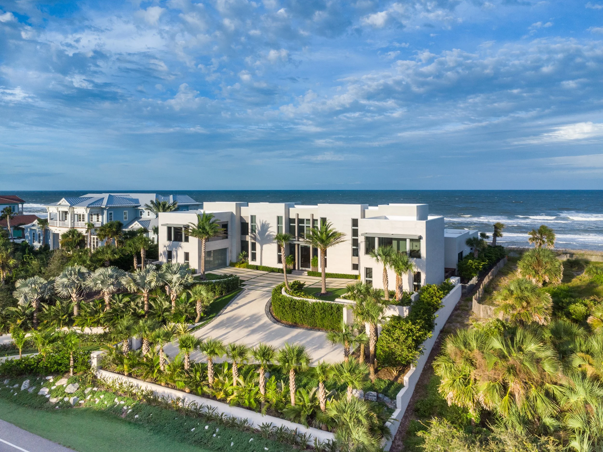 Aerial view of white modern houses with blue roofs and palm trees facing the ocean under a partly cloudy sky.