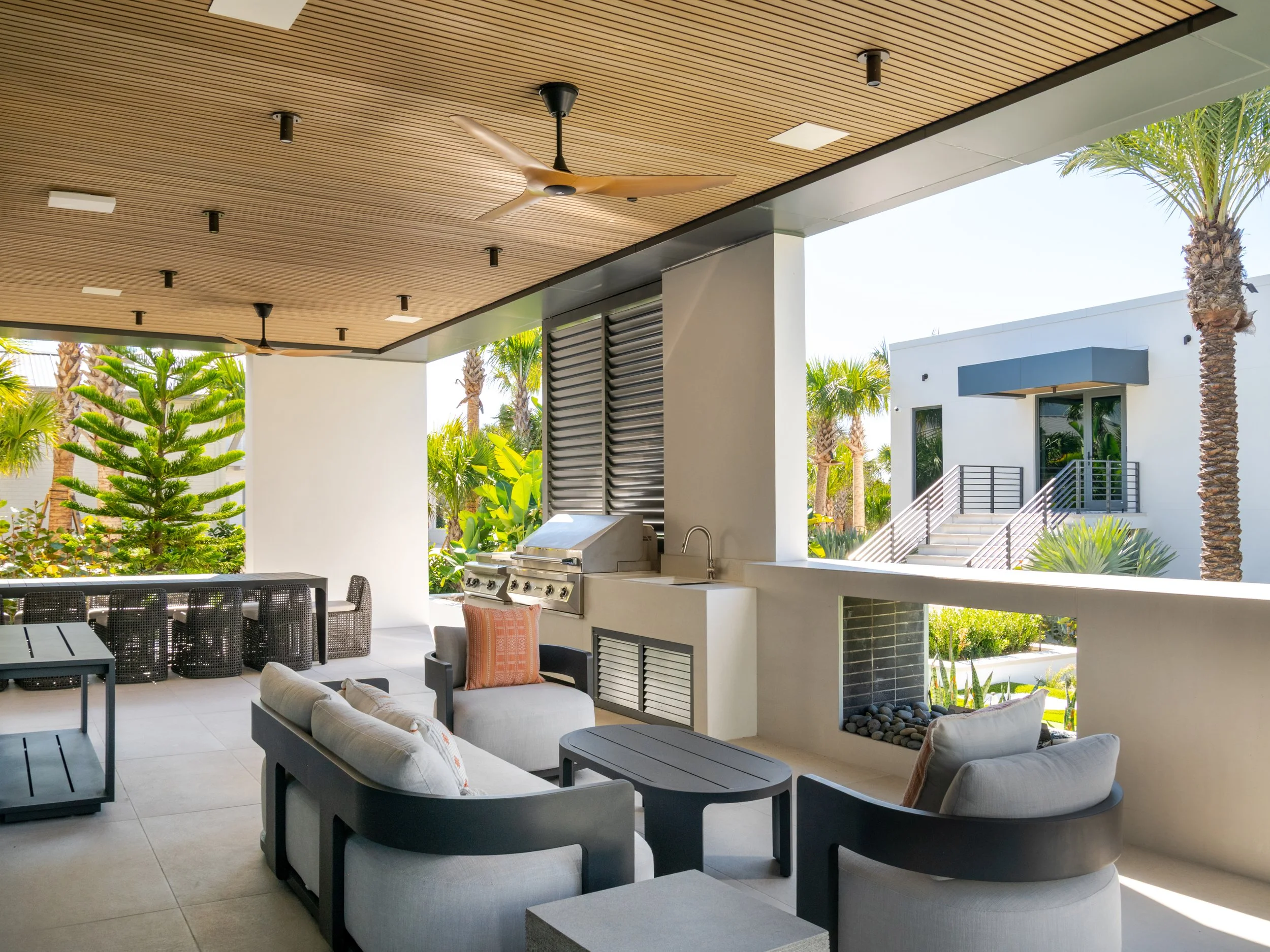 Modern outdoor patio with seating area, ceiling fans, barbecue grill, and surrounding greenery, including palm trees.