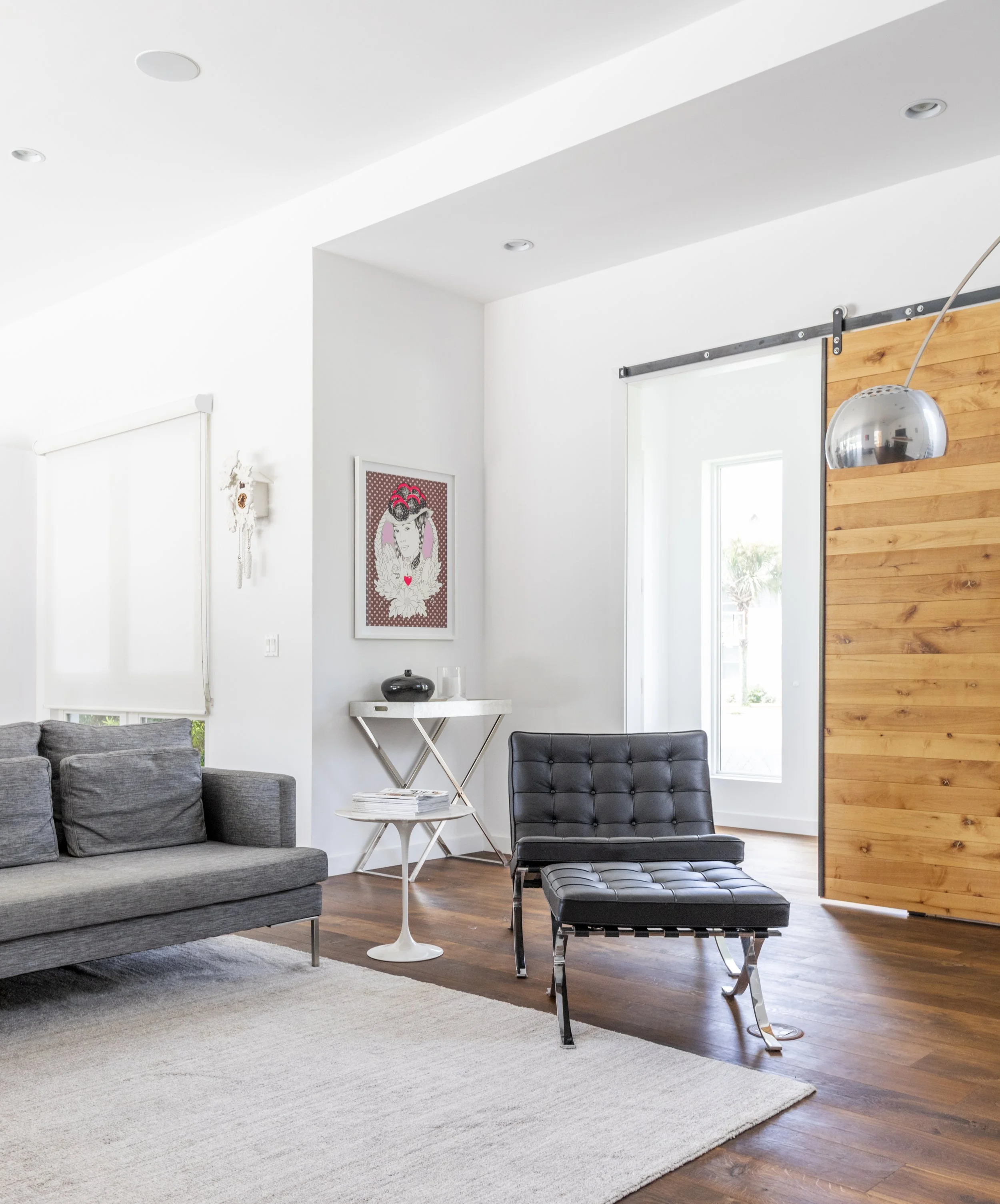 Modern living room with gray sofa, black lounge chair, white side table holding magazines, wall art with a woman, and wooden sliding door, with hardwood floors and a large window.
