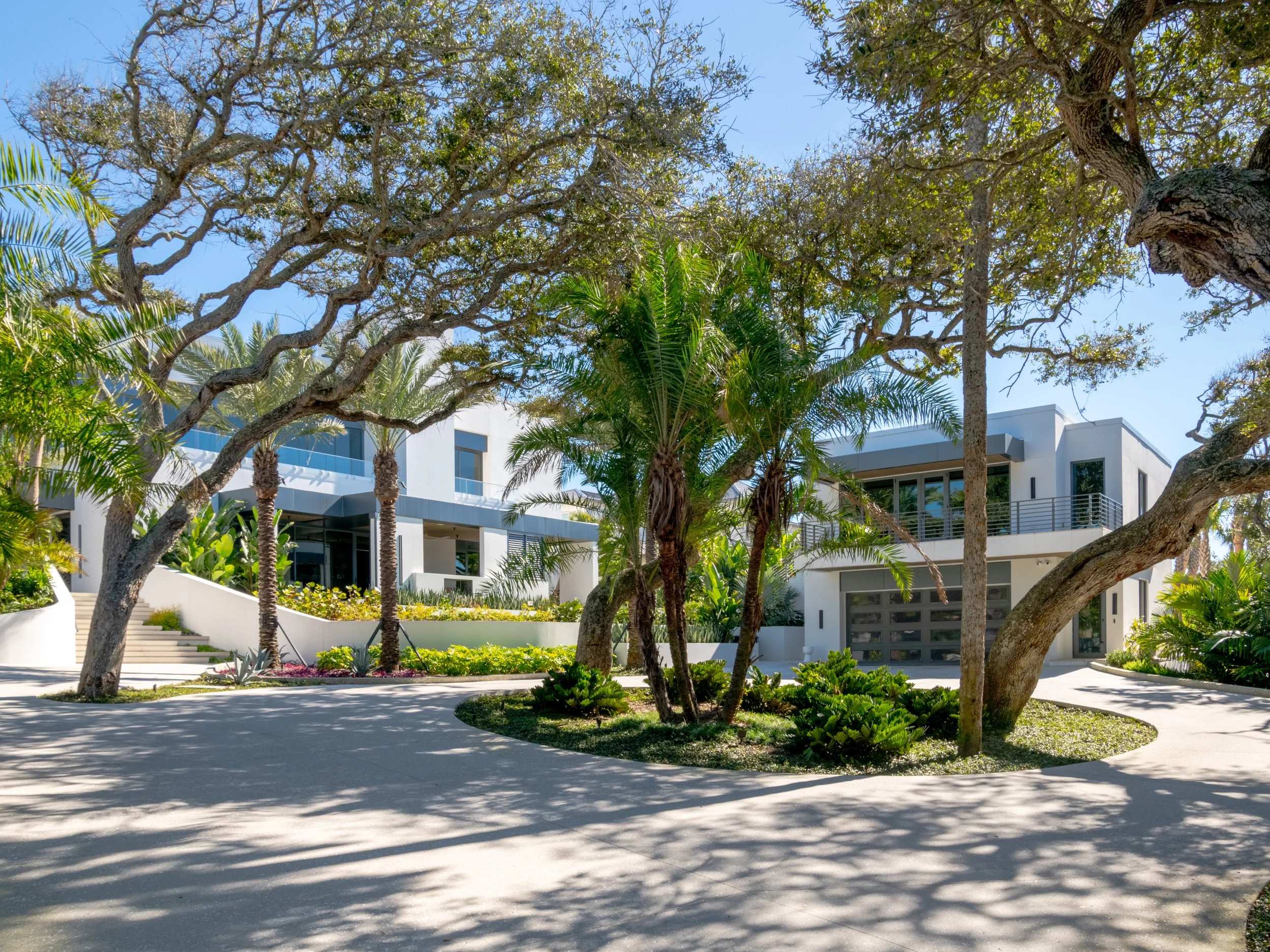 Modern white residential building with multiple floors, balconies, large windows, surrounded by trees, palm trees, and well-maintained landscaping under a clear blue sky.