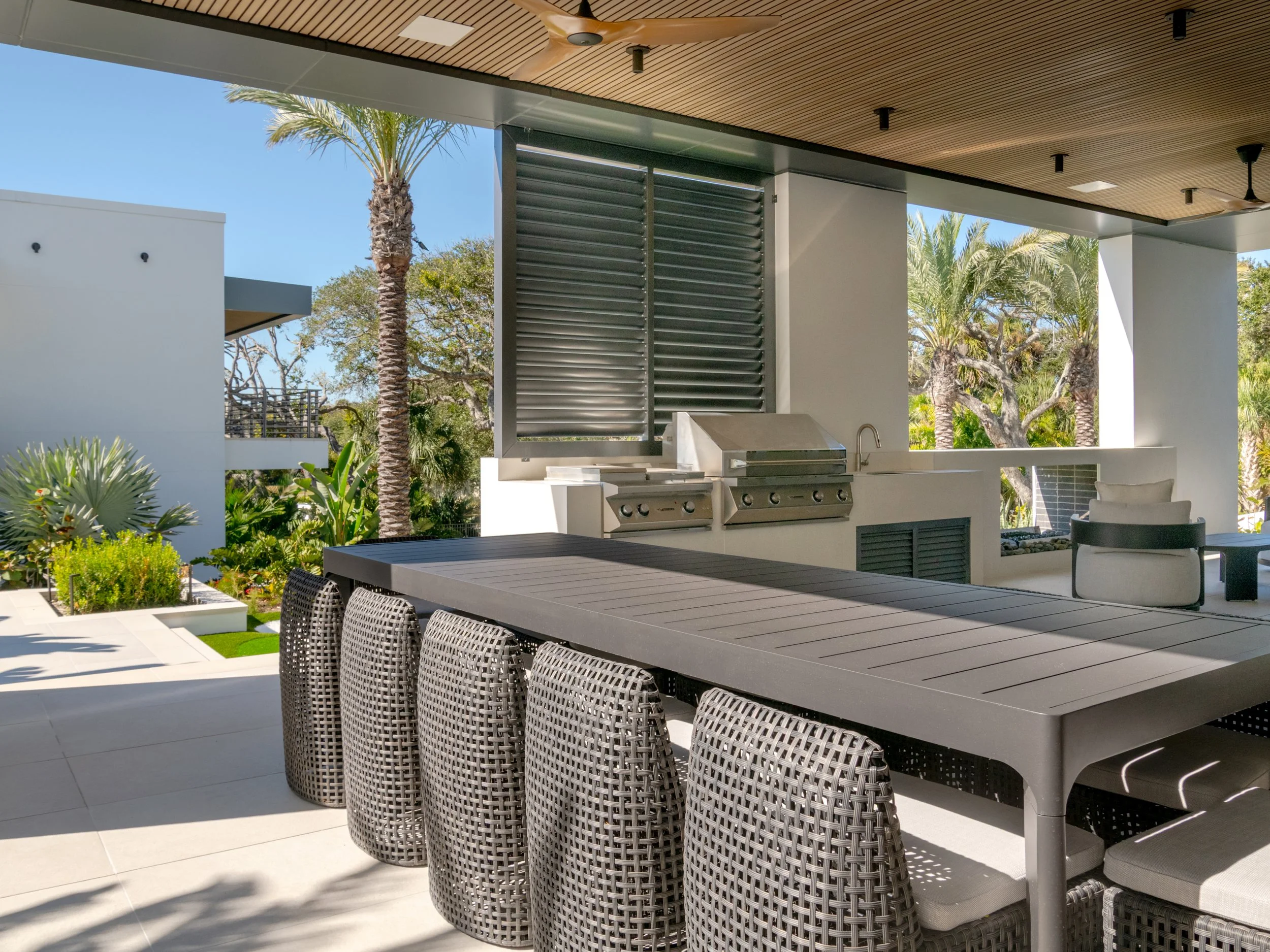 Modern backyard outdoor kitchen with grill, dining table, woven chairs, and surrounding lush greenery including palm trees.