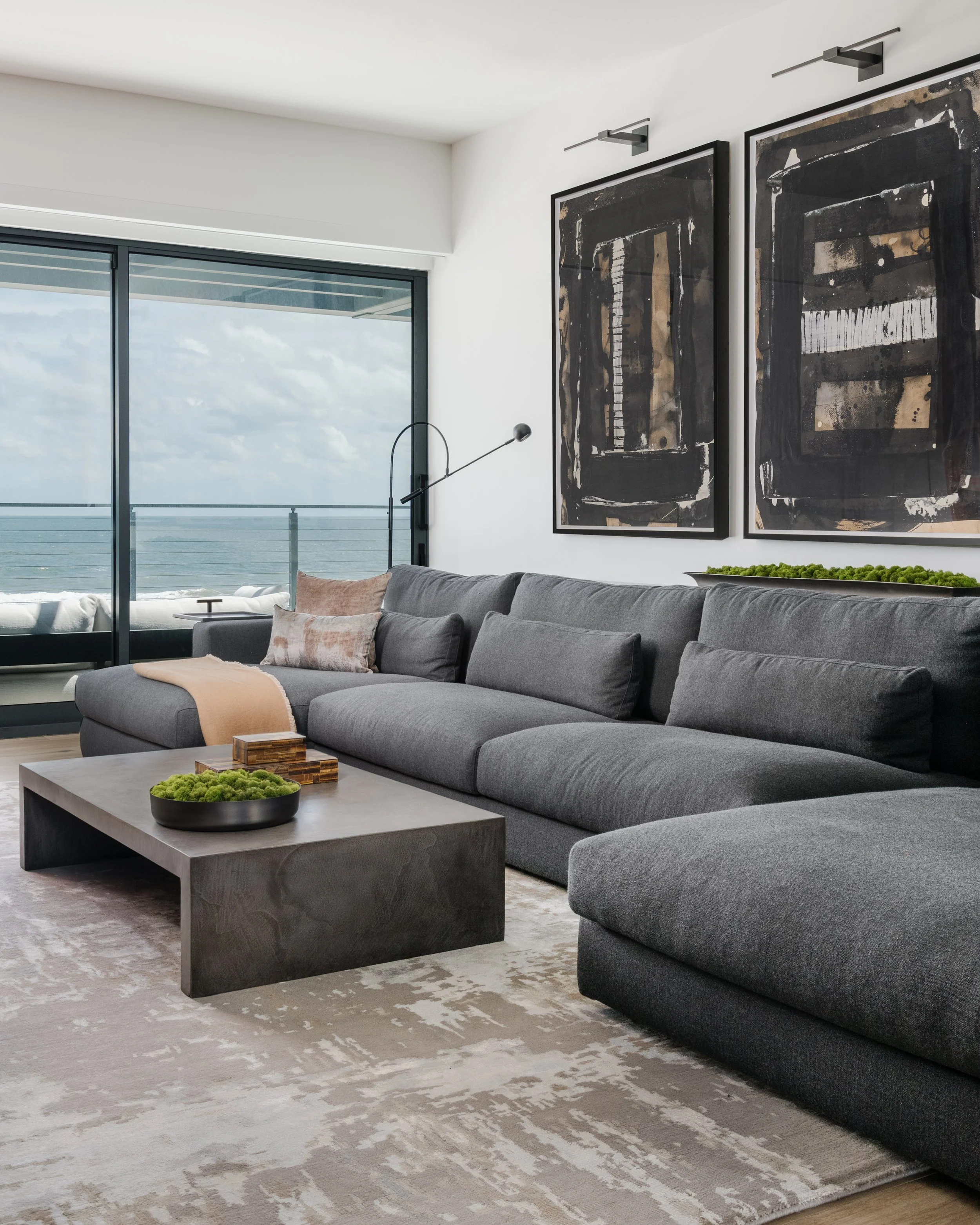 Living room with large sliding glass doors leading to a balcony overlooking the ocean, modern gray sectional sofa with pillows, abstract black and brown artwork on white wall, concrete coffee table with green moss decor, patterned area rug