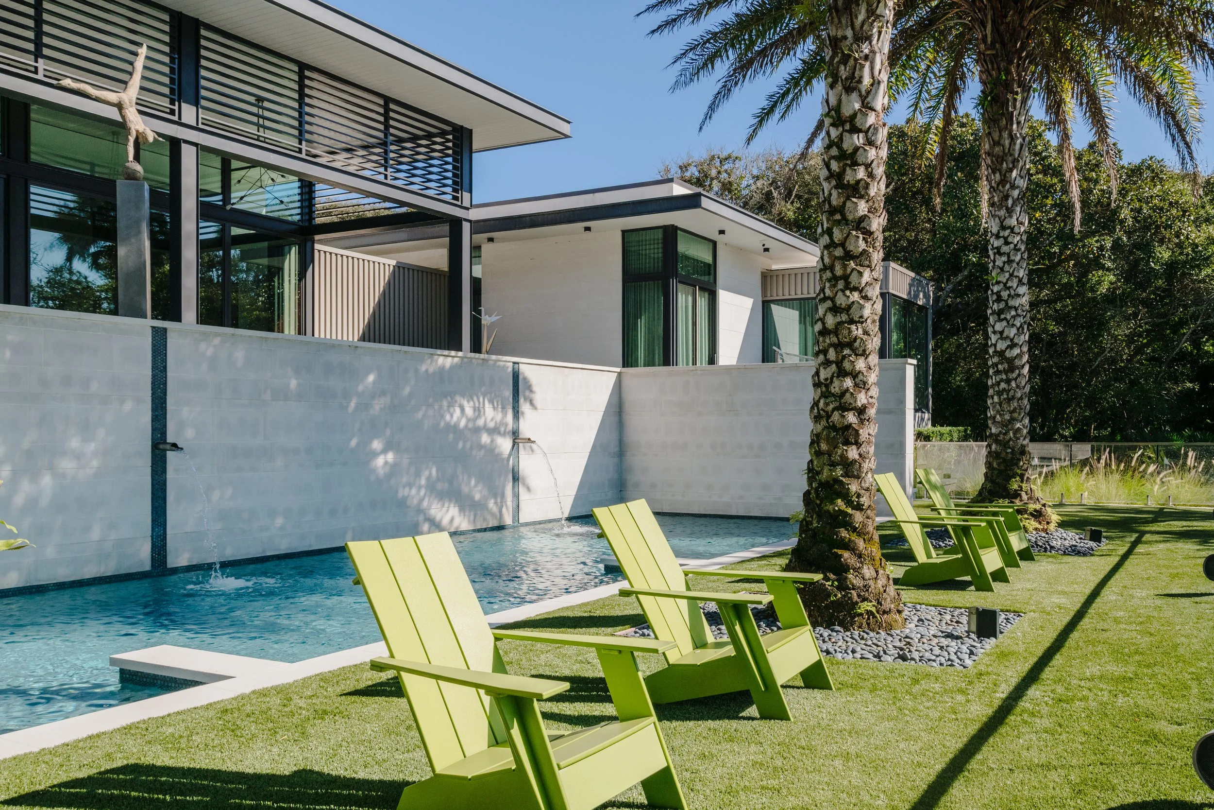 Modern house with large glass windows, palm trees, outdoor pool, and lime green lounge chairs on a grassy area.