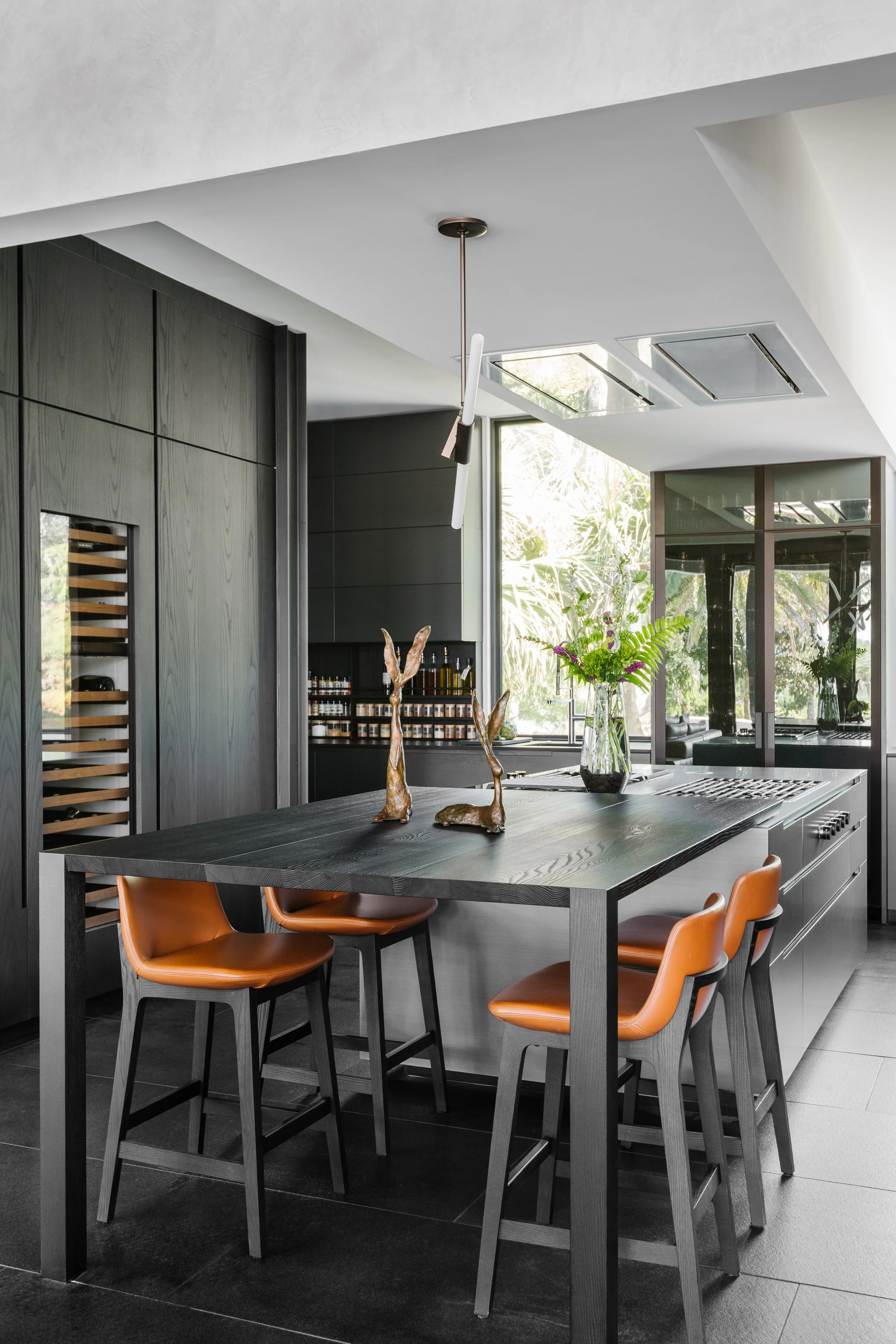 Modern kitchen with black wood cabinets, a black dining table, orange leather chairs, a glass vase with flowers, and a large window with plants outside.