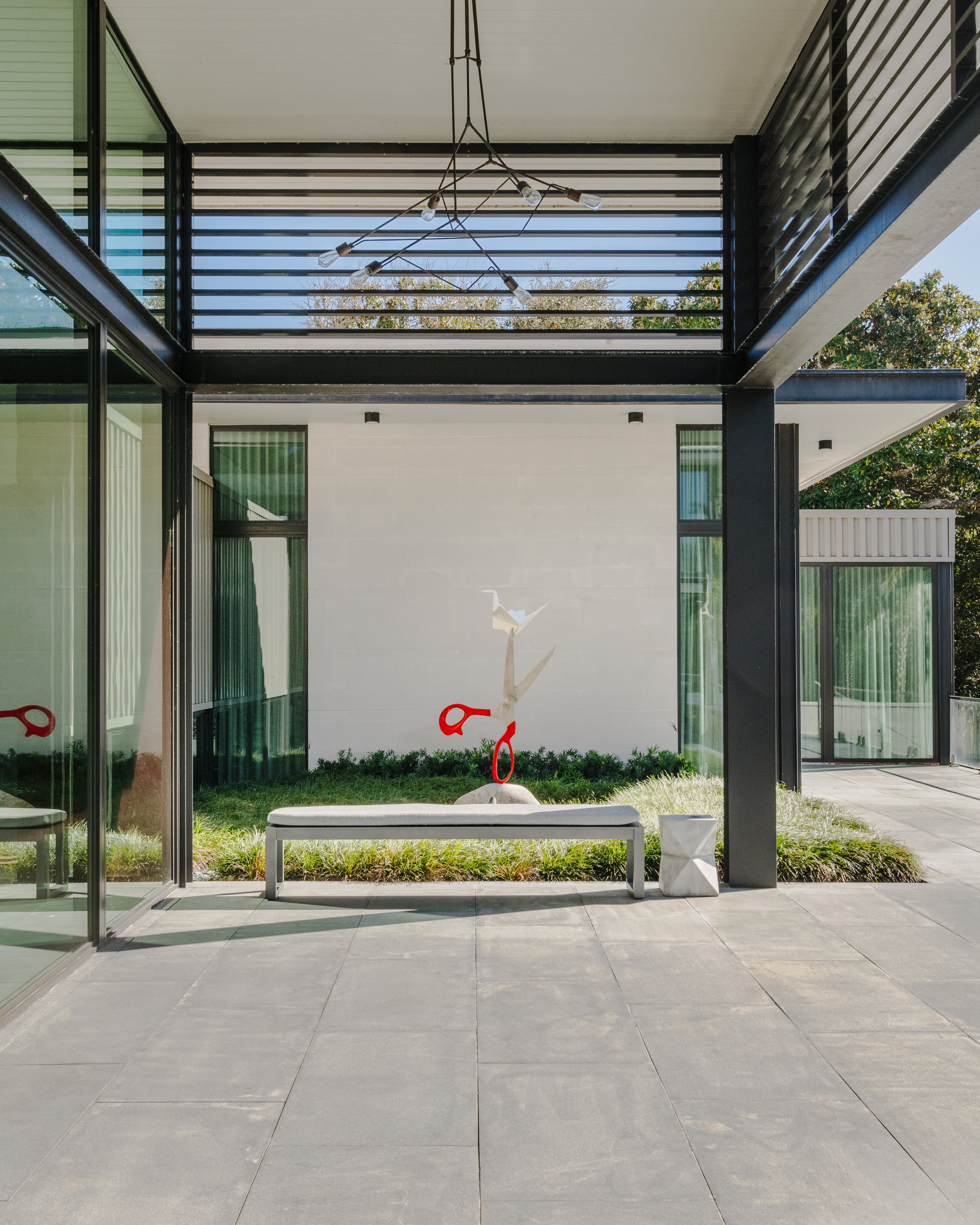 Modern outdoor patio with large grey tiles, glass walls, black metal supports, and a view of a small garden with a white seagull sculpture and green trees.