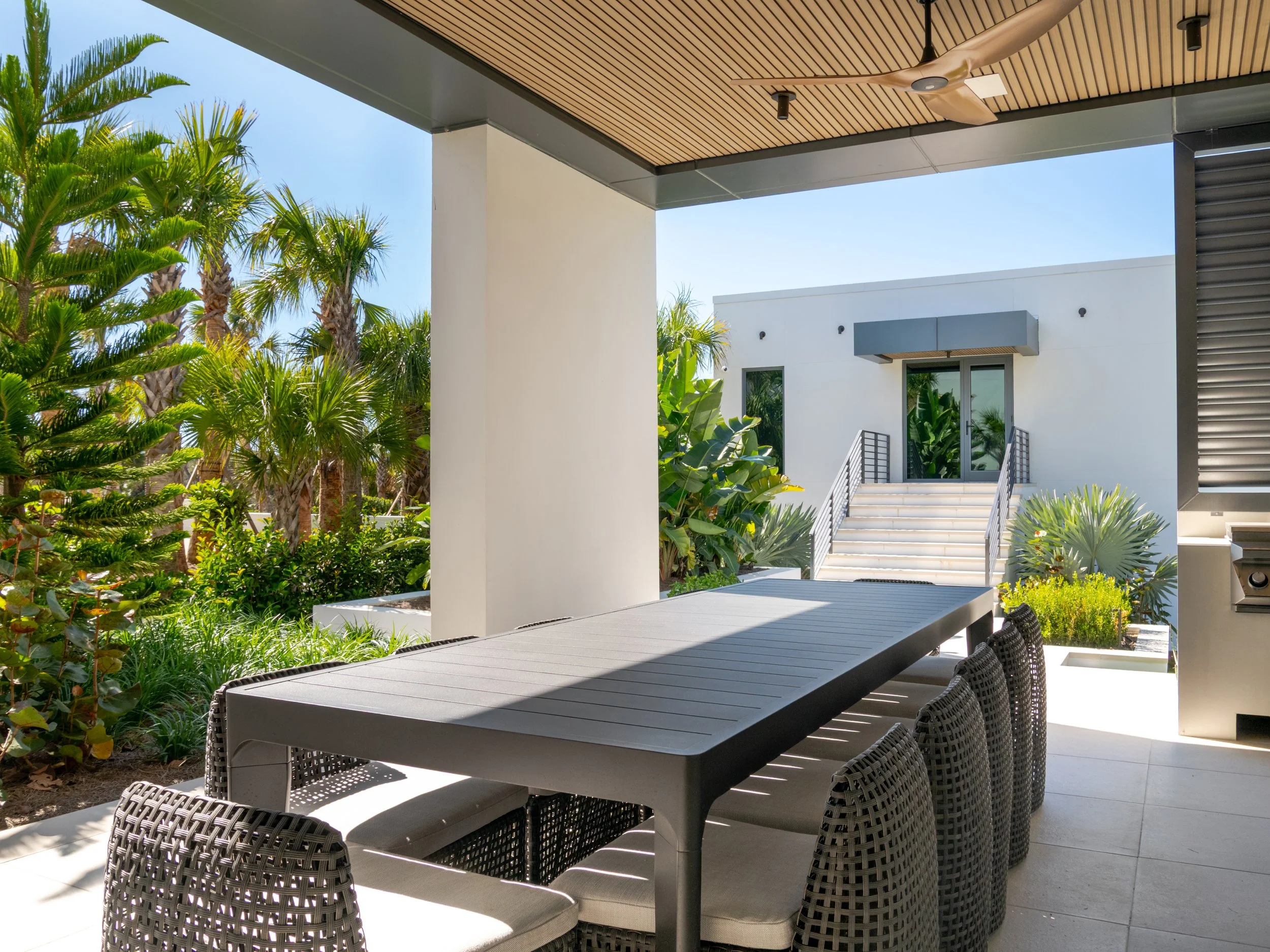 An outdoor patio with a large rectangular dining table and six wicker chairs, surrounded by lush green palm trees and plants, with a modern white building in the background, under a wooden slat ceiling with ceiling fans.