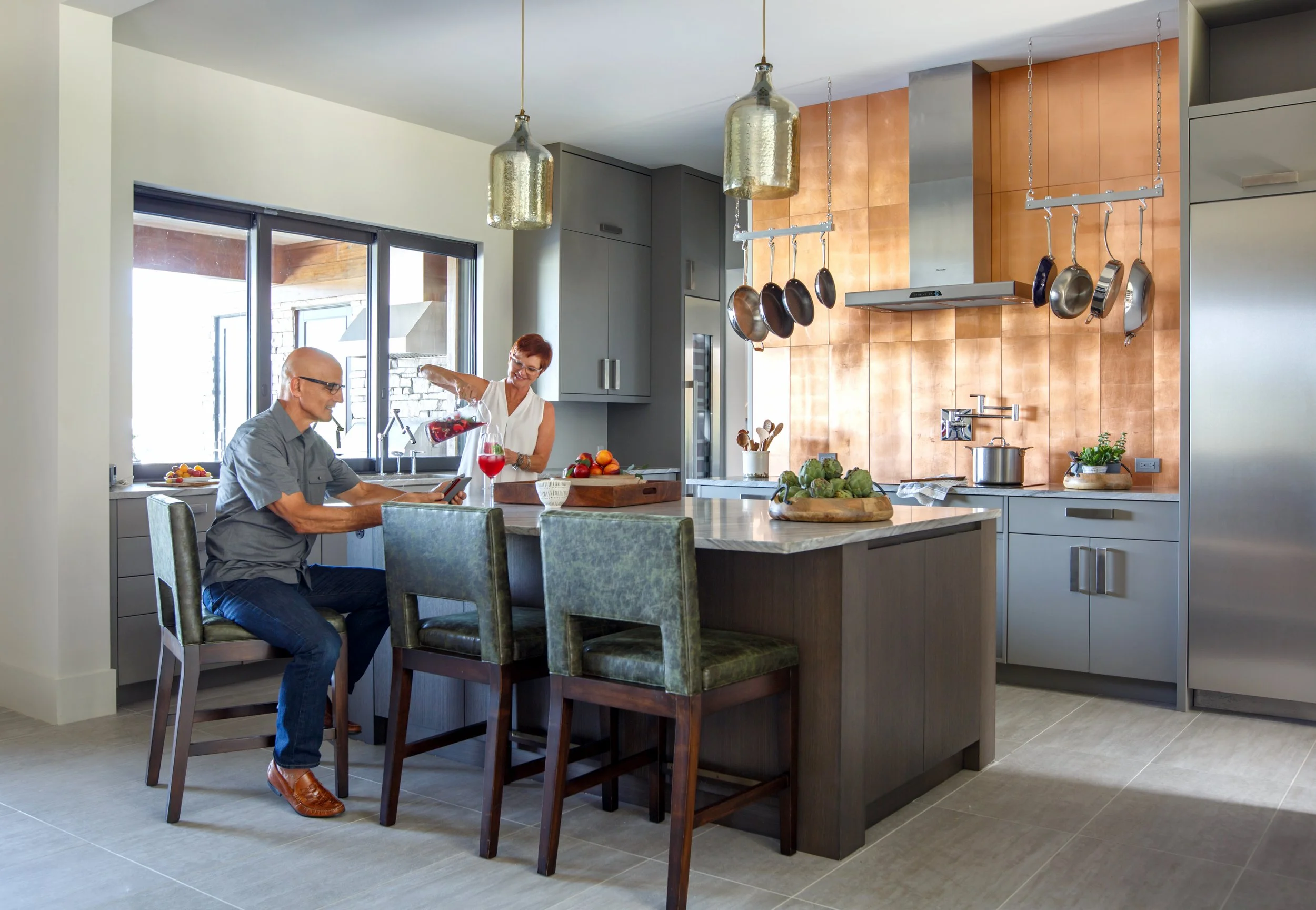 A man and woman in a modern kitchen, the woman is pouring a drink for the man, who is seated at the kitchen island with a tablet. The kitchen features hanging pots and pans, gray cabinets, and large windows.