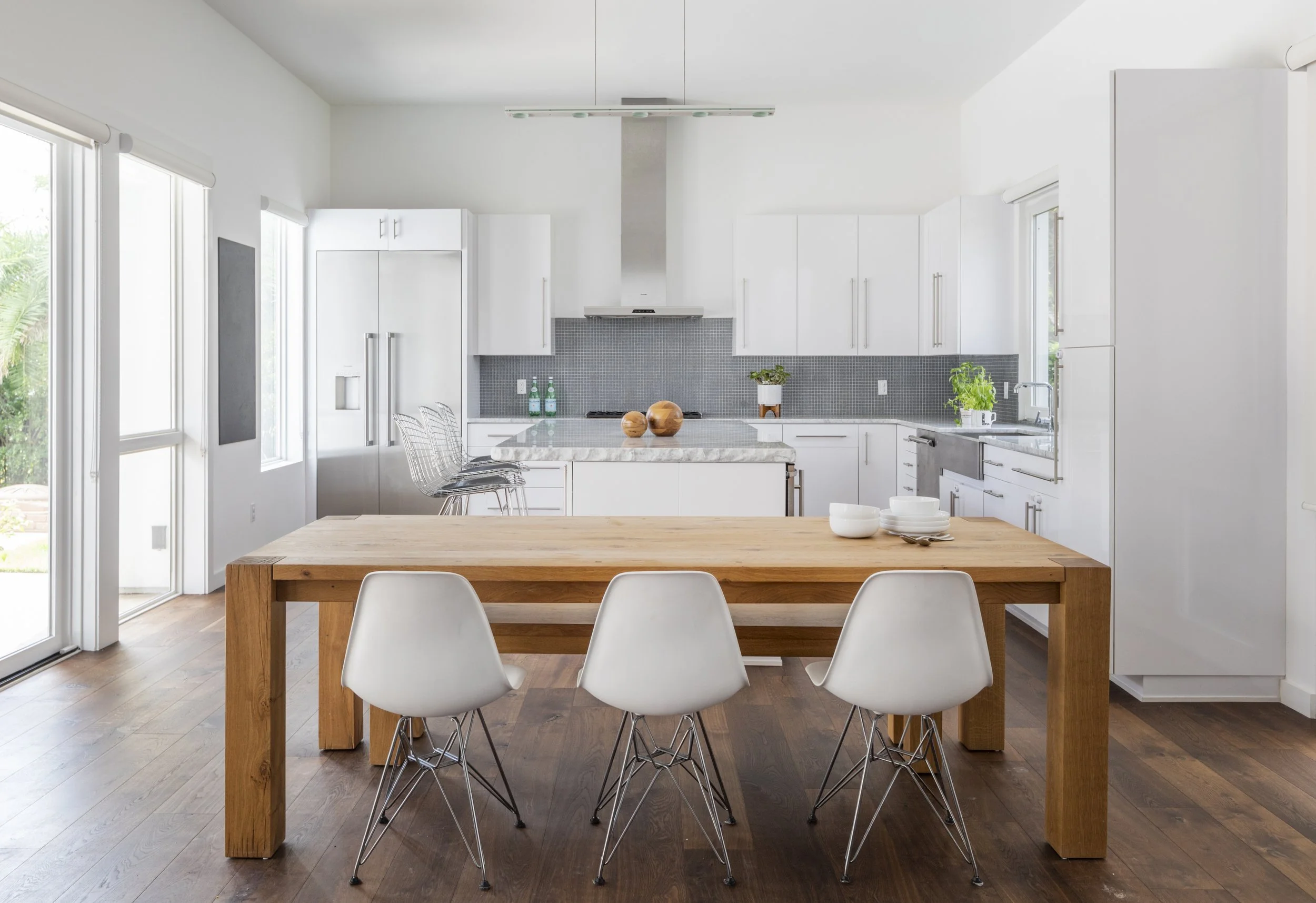 Modern white kitchen with marble island, wooden dining table, and white chairs, large windows, and minimal decor.