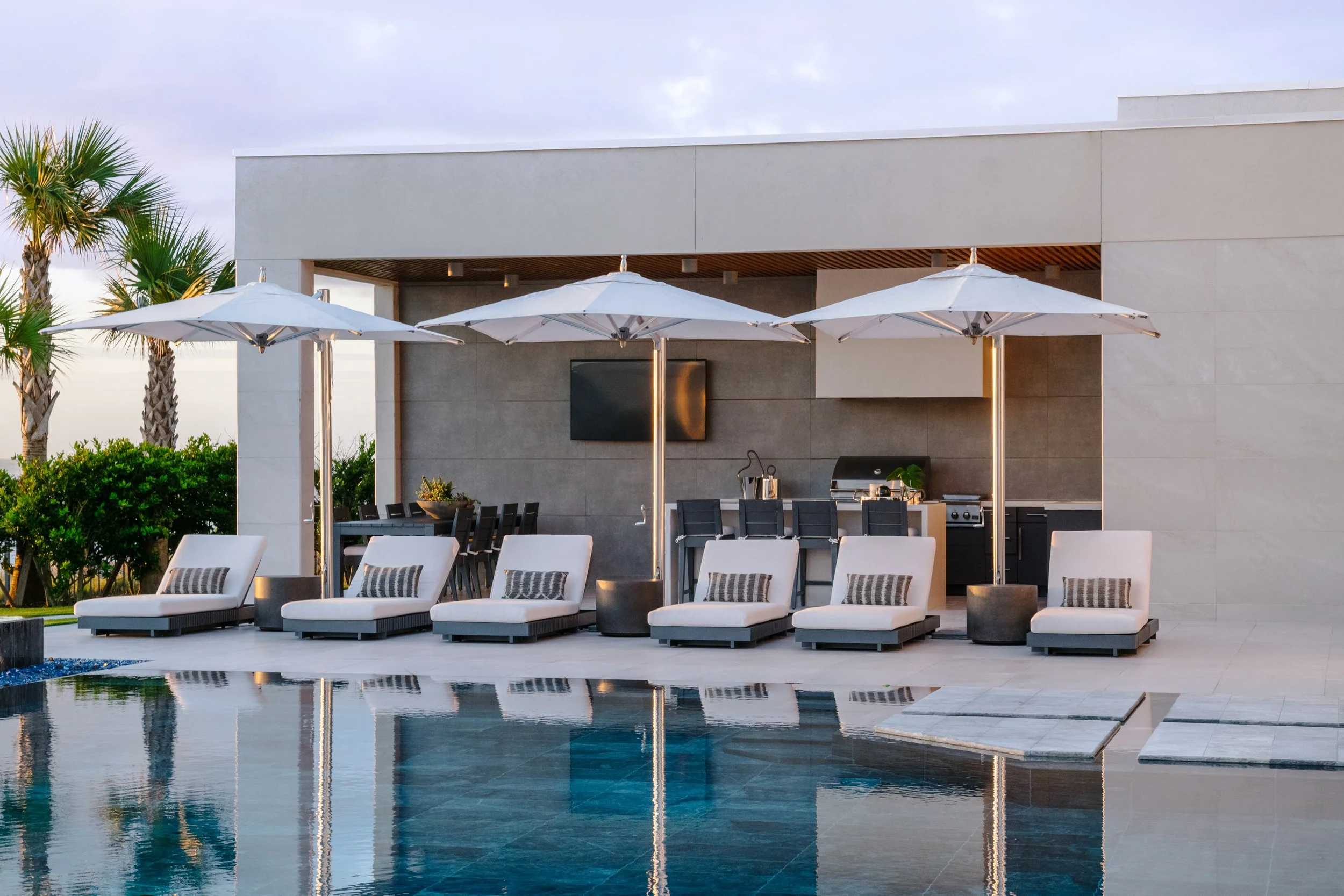 Modern outdoor patio area with white lounge chairs, umbrellas, and a swimming pool, neighboring a contemporary building with palm trees.