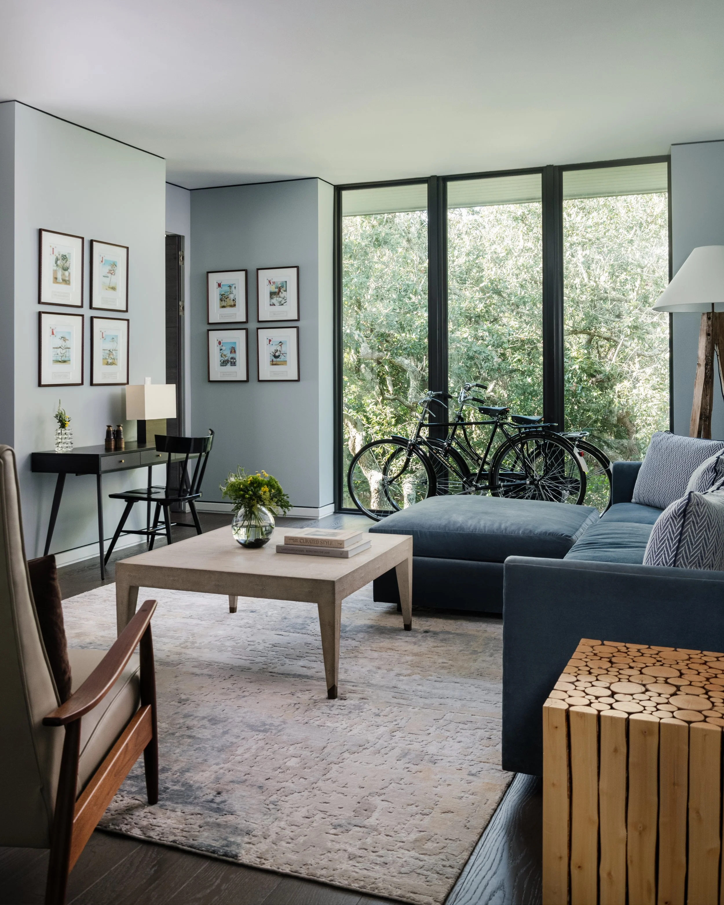 Interior view of a modern living room with large windows showing green trees outside. The room has a blue sofa, a light-colored coffee table with books and a vase of yellow flowers, a patterned rug, and framed artwork on the walls. There are bicycles